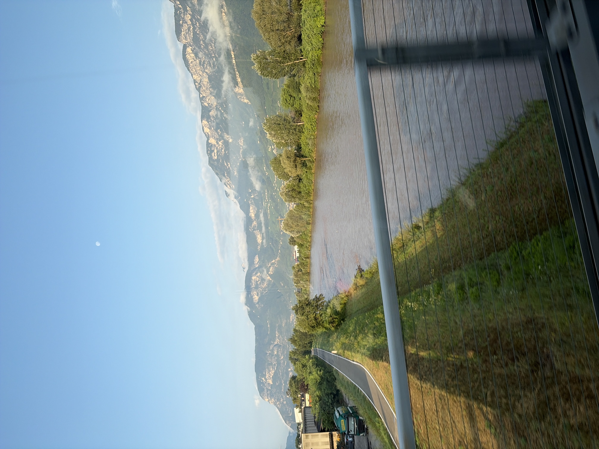 The image depicts a wide, brown river flowing alongside a paved path and a grassy embankment. In the background, a range of rugged mountains rises, partially shrouded in mist and clouds, with the faint outline of the moon visible in the clear blue sky above. On the left side of the riverbank, there are industrial buildings and a green truck. The scene appears to be captured from a bridge, as a metal railing with horizontal wires runs across the foreground. The overall impression is one of a scenic, natural landscape bordering a developed area, likely in Neumarkt - Egna, Italy, given the mountain scenery often associated with the region.