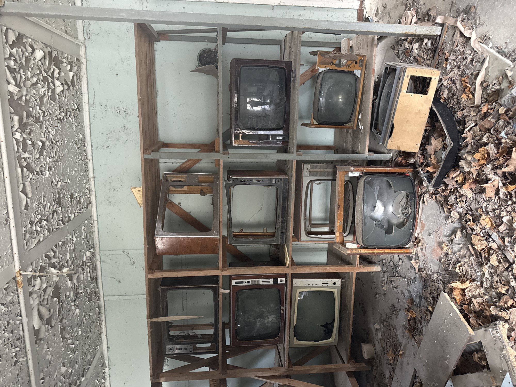 The image shows a dilapidated room in Pryp'yat', Ukraine. A makeshift wooden shelf structure is filled with discarded, old-fashioned television sets, some missing parts or with broken screens. Debris and fallen leaves cover the floor, indicating a state of abandonment and decay. Paint peels from the walls, further emphasizing the deterioration of the environment. There are no people visible in the scene. The objects present include the wooden shelf, several old television sets, and various pieces of debris on the floor. The primary action depicted is the accumulation of decay and neglect within the room.