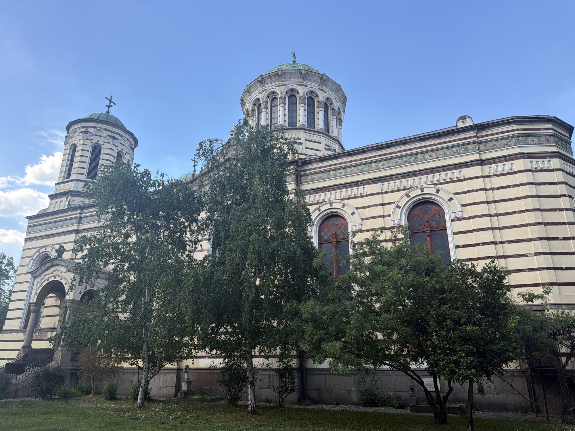 The image shows a full, low-angle view of the Russian Church (officially known as the Church of St. Nicholas the Miracle-Maker) in Sofia, Bulgaria. The church is a cream-colored structure with horizontal stripes of red brick. Its architectural style is characterized by the presence of domes, arched windows, and intricate decorative details. A larger dome is situated atop the main building. A secondary, smaller dome can be seen to the left. Several trees are partially obscuring the lower portion of the building. The sky is clear and blue. The foreground consists of a green lawn. There are no people visible in the image.