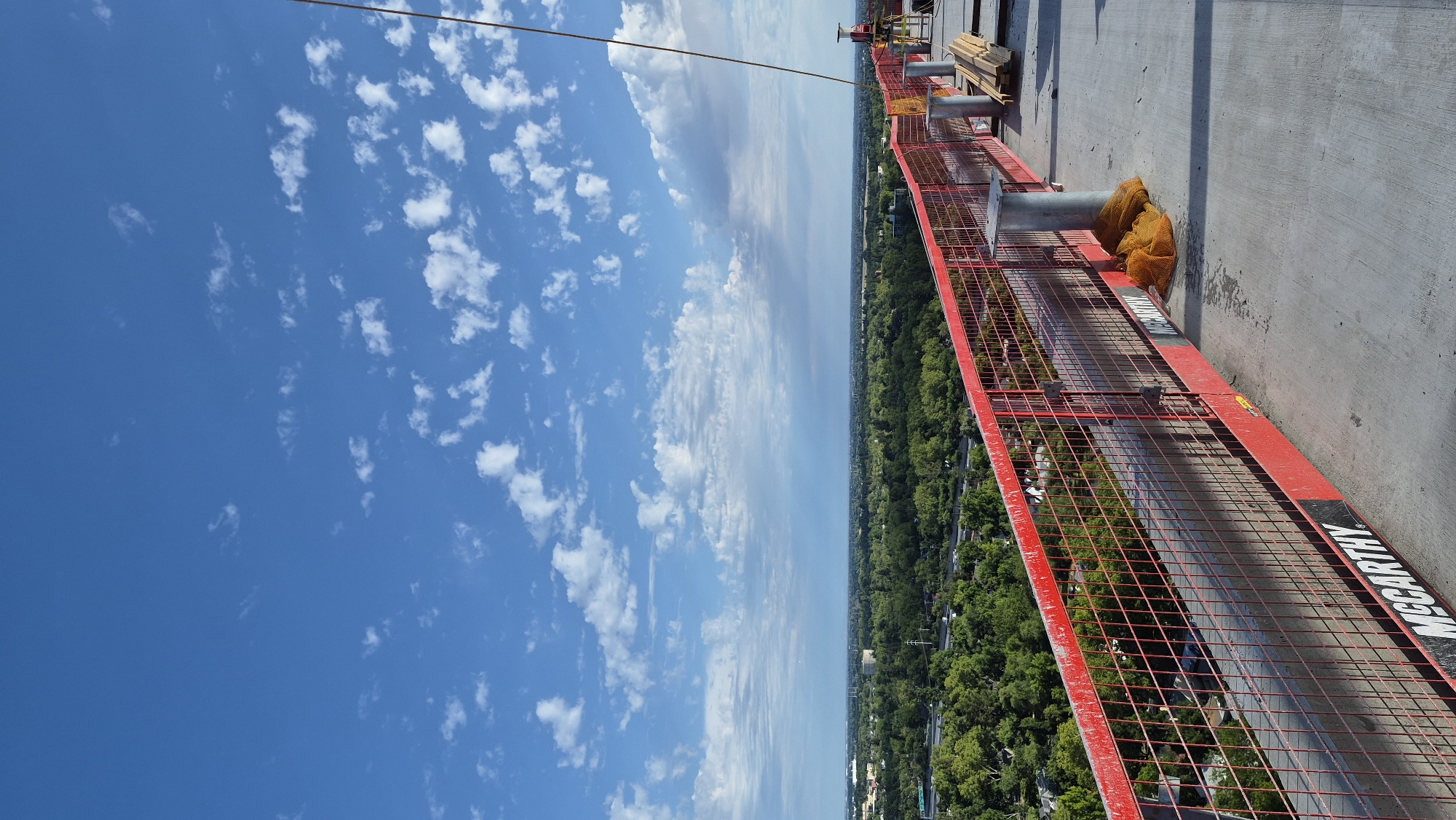 The image depicts a construction site with a concrete floor and a red safety fence. The fence has "MCCARTHY" written on it multiple times. Beyond the fence, there is a lush green landscape with trees and buildings, suggesting a suburban or city outskirts area. The sky is blue with scattered white clouds. In the foreground, there are some construction materials, including wooden planks and orange mesh bags. A thin yellow cable extends from the top right of the frame, likely a safety line. The scene appears to be an elevated perspective, possibly from a building under construction. The location is identified as Sacramento, United States of America.