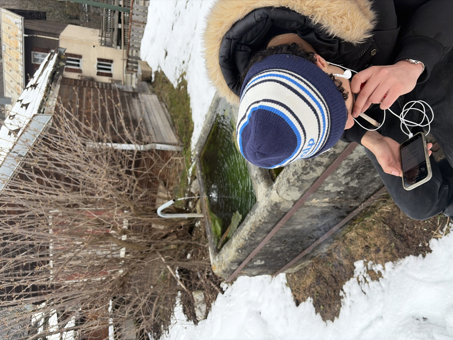 This image captures an outdoor scene in La Clusaz, France, characterized by visible snow patches on the ground, indicative of a winter or early spring season. In the immediate foreground, a person is observed from a slightly elevated, rear-left perspective. This individual wears a dark winter jacket featuring a prominent light-colored faux fur-lined hood and a blue and white striped knit beanie with "alp" visible on it. White wired earphones are present in both ears. The person holds a light-colored smartphone with a dark screen in both hands, positioned to be viewed. Between the thumb and index finger of the right hand, a hand-rolled cigarette or joint is held near the person's mouth.
In the midground, a rectangular, weathered grey stone water trough is centrally positioned. It contains green-tinted water, suggesting the presence of algae, into which a curved, silver-colored metal spigot continuously dispenses water, creating concentric ripples. The near side of the trough features two horizontal, rust-colored metal bars.
The background consists of a large, dense deciduous bush with bare branches situated behind the water trough. To the left and slightly behind the bush, a rustic wooden building with dark horizontal planks and a light-colored roof is visible. Further into the background, to the right, another multi-story building with a light-colored facade and dark window shutters stands on elevated terrain. The ground around the trough and extending into the background shows mixed areas of snow and exposed brown earth or dead vegetation.