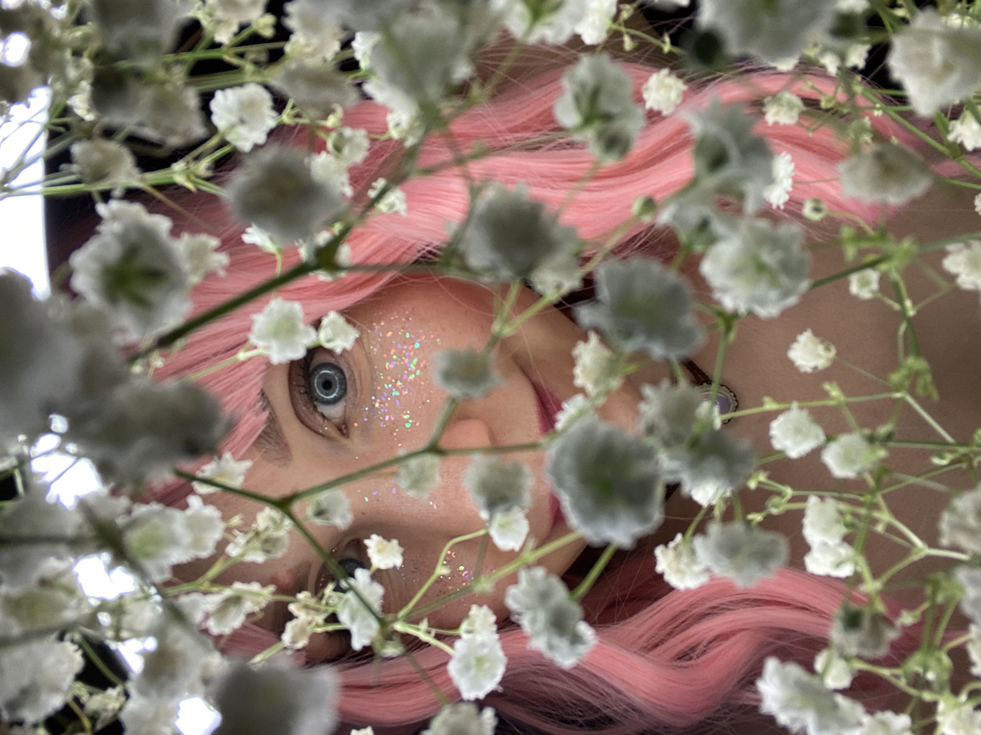 The image is a close-up, high-angle portrait featuring a person partially obscured by a dense foreground of white, delicate flowers, resembling baby's breath (Gypsophila). The perspective is from above, looking down through the flowers, and the subject appears to be rotated sideways, with their head slightly tilted.
The primary subject is a person with vibrant, wavy pink hair, which cascades around their head. Only parts of their face are clearly visible through gaps in the foliage. Their eye, on the left side of the frame, is a striking blue or grey color, and appears wide open, looking towards the viewer. Glitter in iridescent or multi-colored flakes is applied to the cheekbone area below this eye, adding a whimsical or artistic touch. A subtle hint of the person's mouth and chin is visible towards the bottom right.
The small white flowers in the foreground are largely out of focus, creating a soft, blurred effect that frames the subject's face. The background, behind the person's head, appears dark and indistinct. There are no discernible location cues within the image, and no visible text. The lighting is soft, highlighting the colors of the hair, eyes, and glitter. The overall mood of the image is ethereal and artistic.