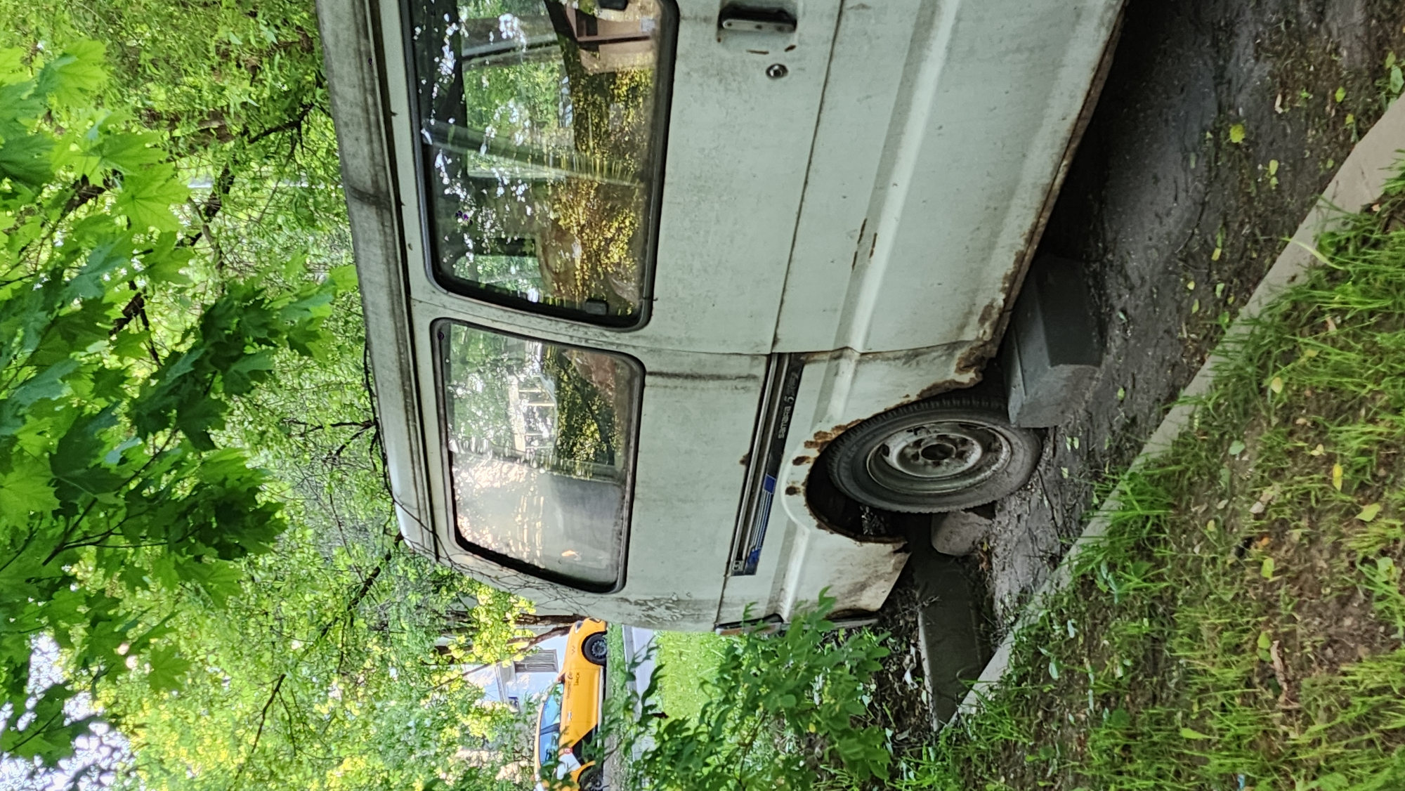 A white, rusty van is parked on the side of a road. The passenger side of the van is visible. The van has two windows and a side door. The tire on the passenger side is visible. A yellow car is partially visible behind the van. Green foliage surrounds the van. The scene is in Moscow, Russia, as indicated by the Cyrillic writing on the side of the van that reads "Samara 5."