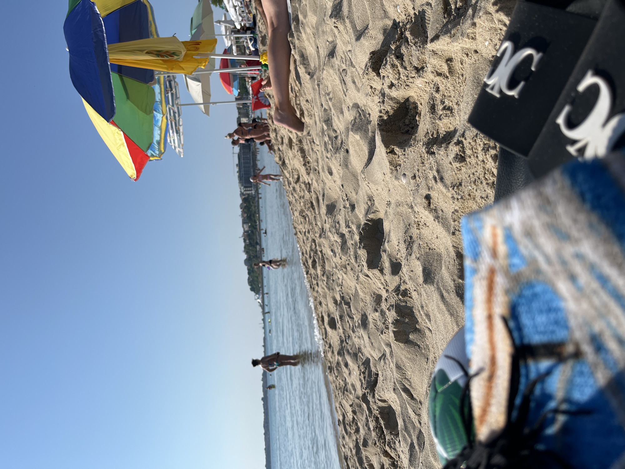This image depicts a sunny day at the beach in Nesebar, Bulgaria. The foreground is dominated by rippled sand, with a partially visible towel and two black objects with white "CC" logos in the lower right corner. In the middle ground, several people are seen wading in the calm sea, and a child is running towards the water. Beach umbrellas, predominantly colorful ones, are set up on the sand. The background shows a coastline with buildings and more umbrellas, under a clear blue sky. The sea is calm and reflects the bright sunlight.