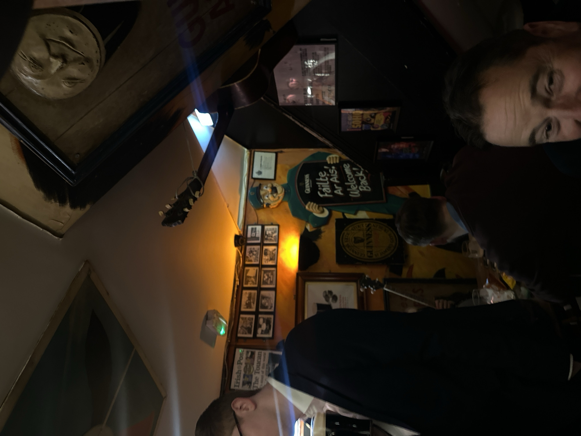 The scene appears to be inside an Irish pub, possibly in London, United Kingdom, based on the decor and signage. The image is taken at an angle, looking upwards toward the ceiling and the walls of the pub. On the walls, there are framed pictures, including one with a face on it, and various Guinness-related posters and signs. One sign reads "Guinness Fáilte Ar Ais! Welcome Back!". There is also a series of smaller framed pictures, potentially showing historical images related to the pub or the surrounding area. Two men are visible, one is partially obscured and another is looking at the camera. One man is wearing a dark suit. There is also part of a guitar in the frame. The lighting is somewhat dim and warm, creating a cozy atmosphere.