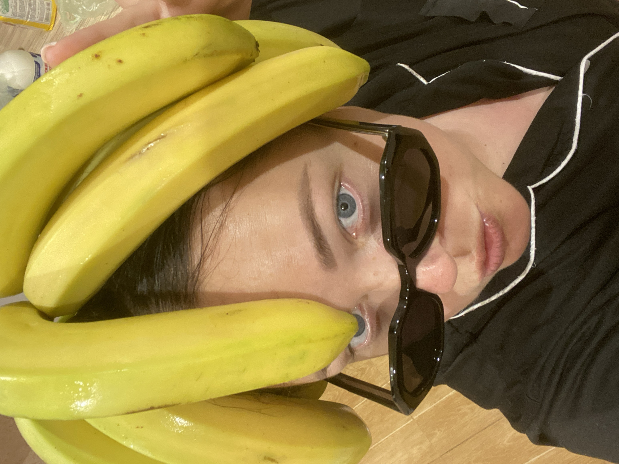 A person with light skin, light blue eyes, and dark hair is lying on their back on a light brown wooden floor. They are wearing black, square-framed sunglasses and a black top with white piping. A cluster of yellow bananas is positioned over their head and partially covering their face. The location is not discernible.