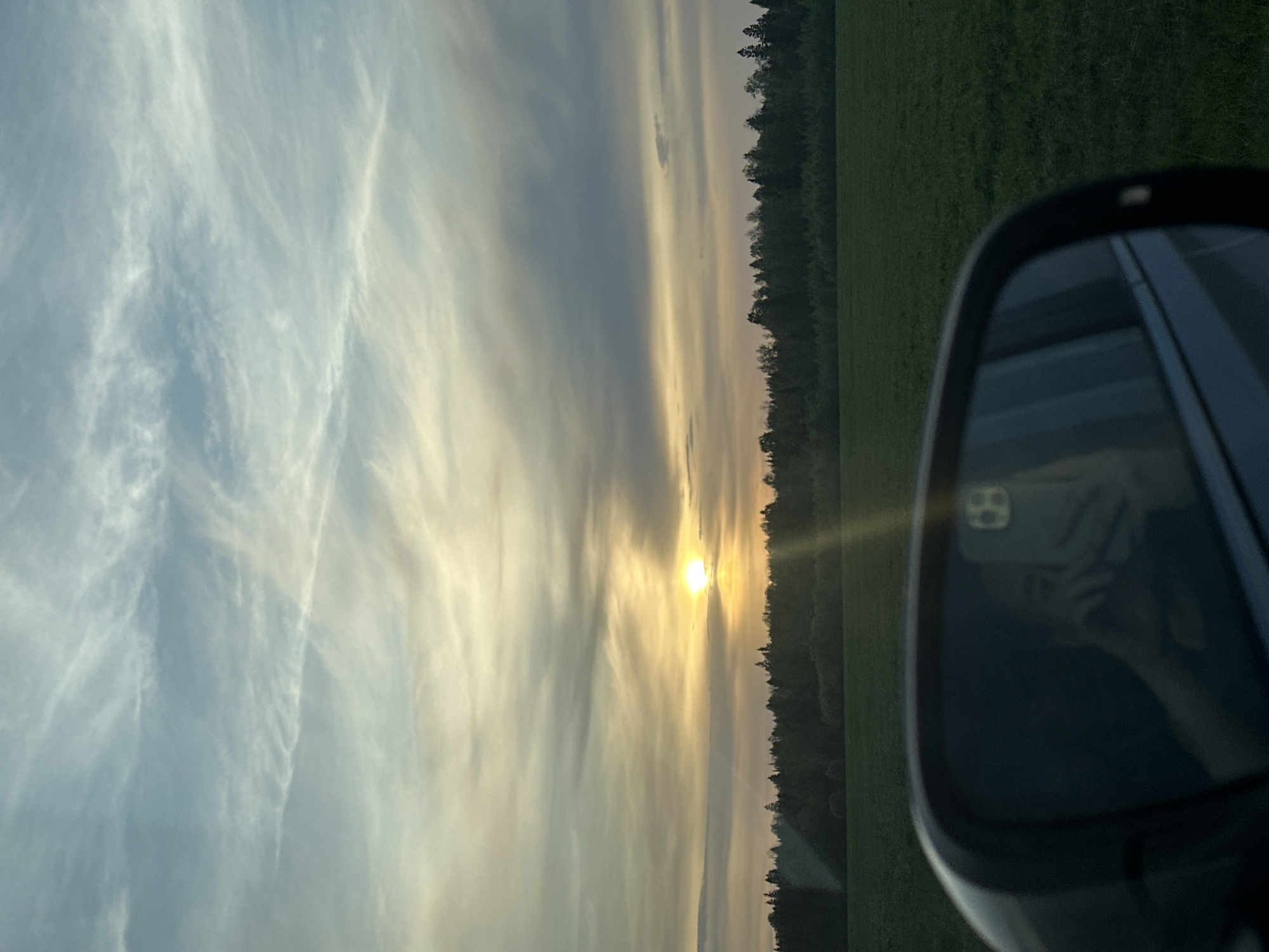 The image depicts a landscape during either sunrise or sunset, viewed partially through the side mirror of a car. The sky is filled with layered clouds, with the sun appearing low on the horizon, emitting a golden glow. To the right, a field or meadow extends towards a line of trees. The car's side mirror is visible in the foreground, reflecting a person holding a phone. The location cannot be definitively determined as Postol, Russia, from the visual information alone.