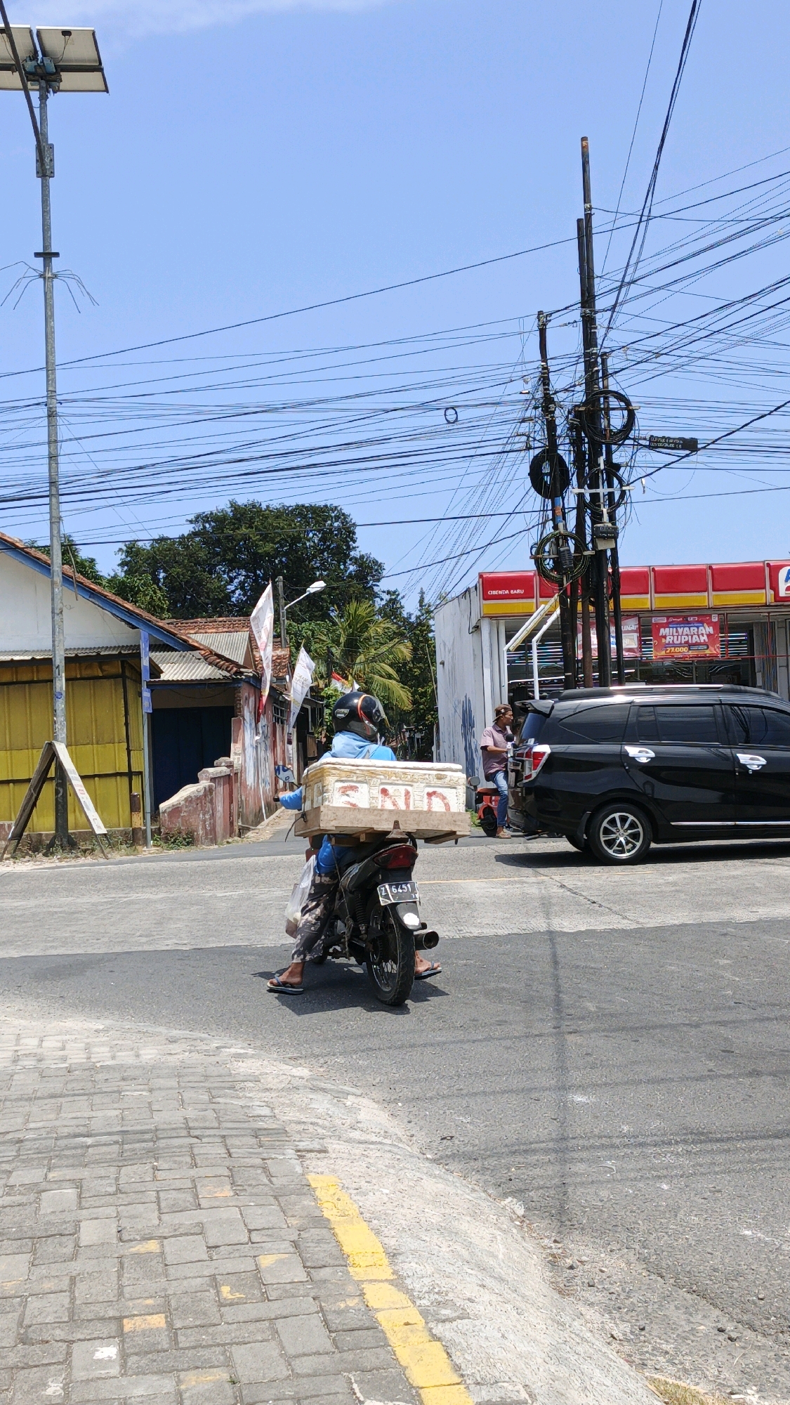 A medium shot captures a street scene in Cibenda, Indonesia on a bright, sunny day, with a clear blue sky. A person wearing a blue long-sleeved shirt and patterned pants rides a black motorcycle. They are wearing a black helmet with a silver visor and flip-flops. The motorcycle carries a large, rectangular, wooden box strapped to the back. The box appears to have some writing on it, possibly "SND". The motorcycle's license plate is partially visible, reading "Z 6451".
In the background, a street intersects, with buildings and a large utility pole laden with numerous black electrical wires dominating the scene. To the left, a yellow building with a blue doorway is visible, next to a smaller building with a red roof. Further to the right, a red and yellow building, likely a storefront or gas station, is present. A black SUV is parked on the right side of the street, and another person is standing near a red scooter. The road is paved, with a brick sidewalk to the left and a yellow line marking the edge of the road. The overall impression is a snapshot of daily life in a busy urban environment.
