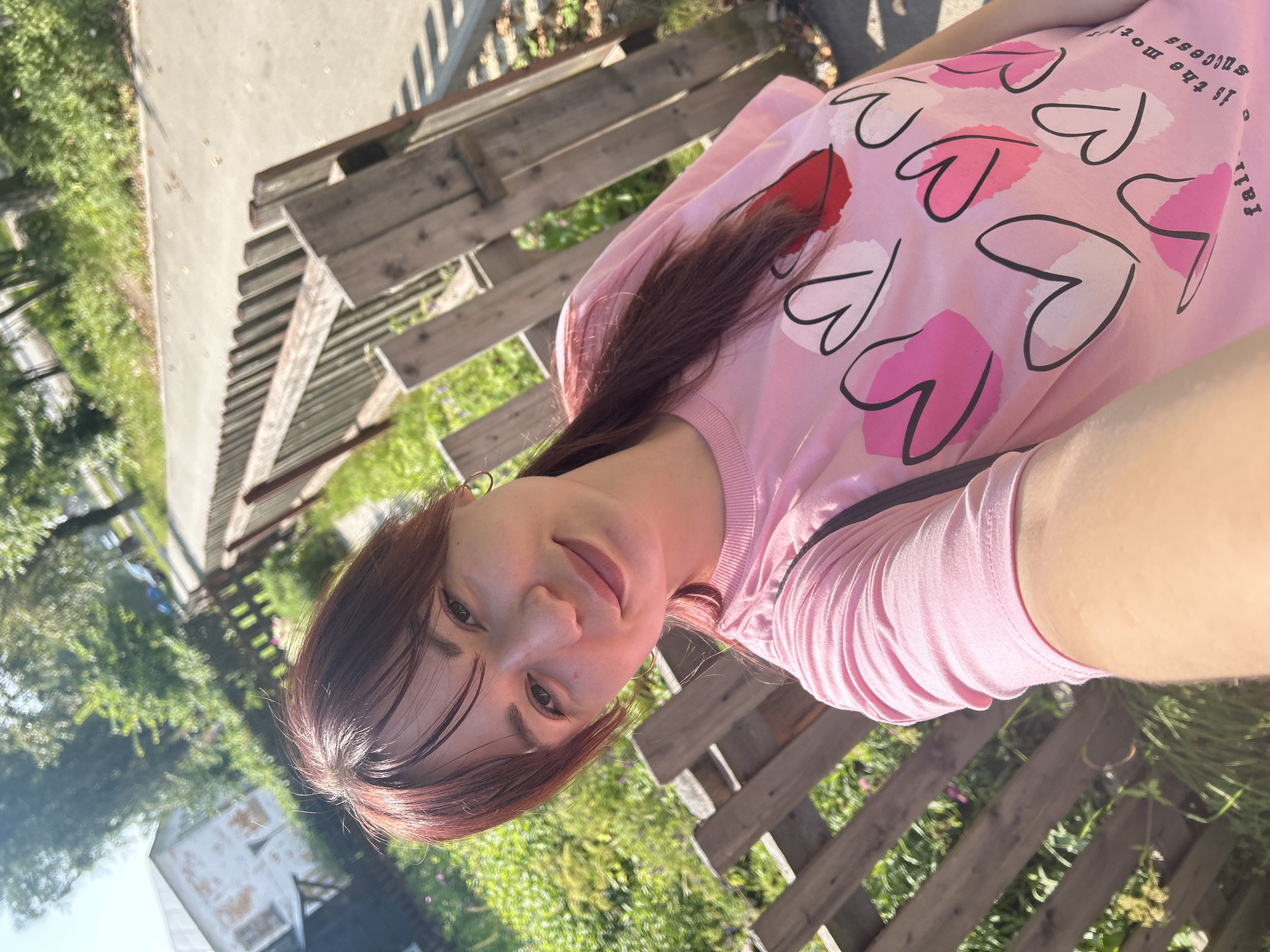 A young woman with shoulder-length reddish-brown hair, wearing a pink t-shirt with a heart pattern, is taking a selfie outdoors. She is smiling and looking at the camera. Behind her, a wooden fence runs along a paved path. Green vegetation and a building are visible in the background. The overall scene suggests a casual outdoor setting, possibly a park or residential area in Krasnovishersk, Russia.