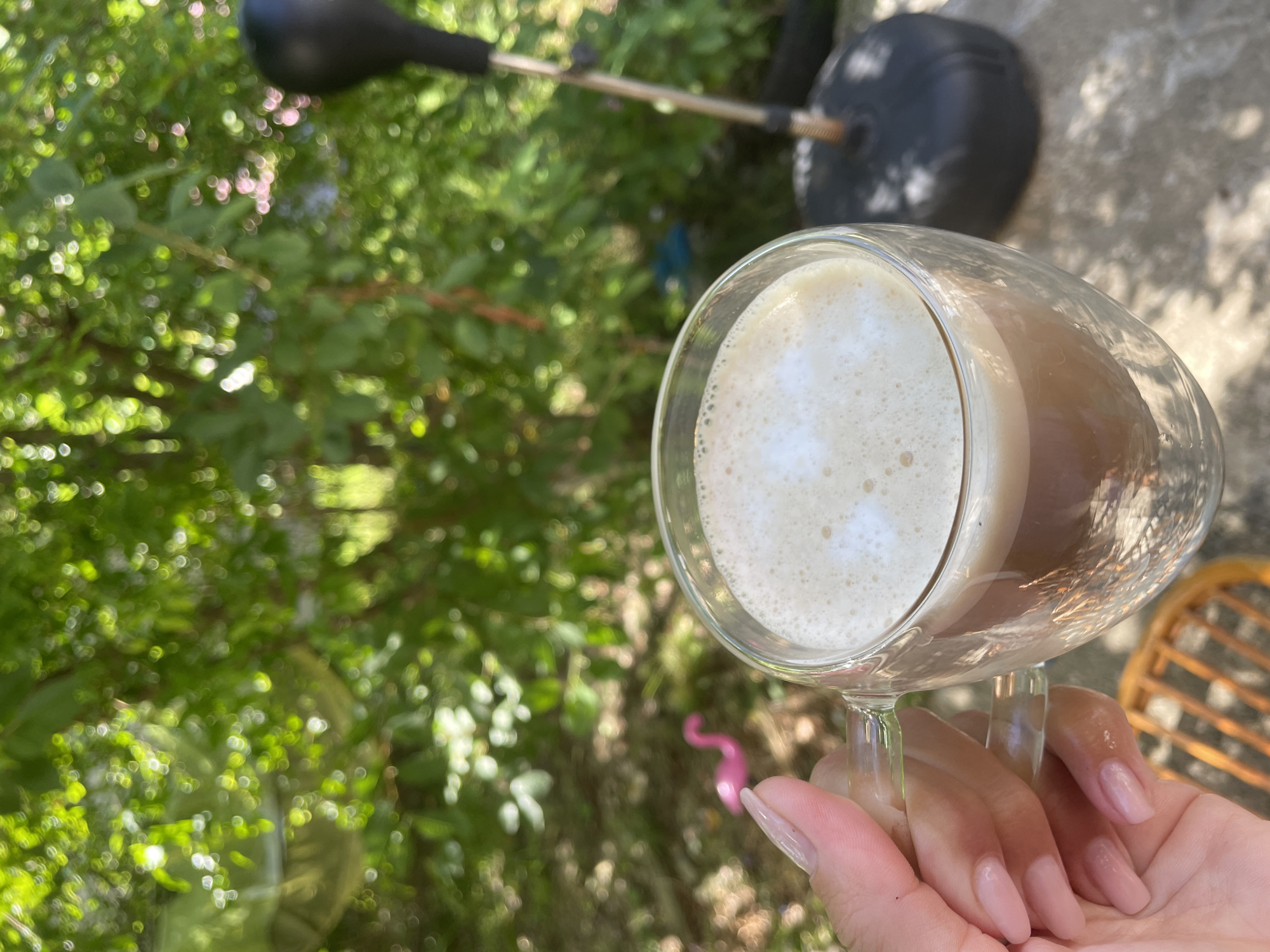 In Baranovka, Russia, a hand with manicured nails holds a clear, double-walled glass mug filled with a frothy, light brown beverage, likely coffee or tea, outdoors. The background is a blur of green foliage, suggesting a garden or park setting, with sunlight filtering through the leaves. To the left, a small pink flamingo figurine is visible among the plants. To the right, a black punching bag on a stand is partially in view, along with a wooden chair in the foreground. The overall impression is a relaxed, casual moment outdoors.