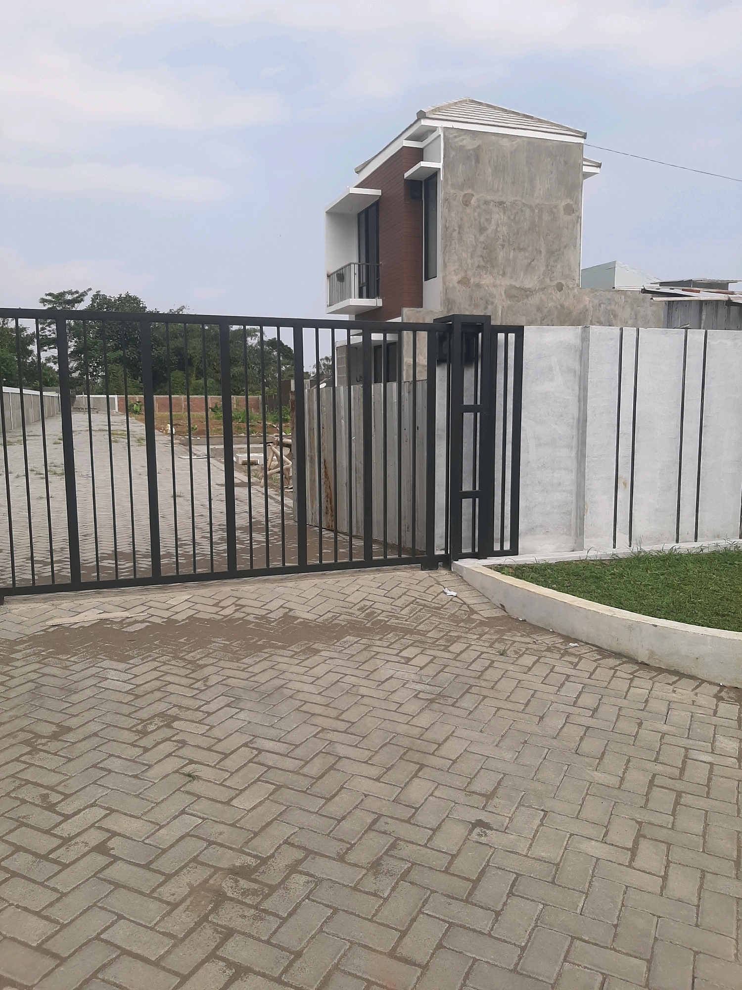 The image shows an outdoor scene with a modern house under construction and a black metal gate. The house has a grey concrete exterior, a brown wooden facade, and white trim. It also features a balcony and large windows. The gate is tall and spans across a paved driveway made of interlocking bricks. Behind the gate, the ground appears to be under development with some construction materials visible. To the right of the gate, a concrete wall runs along the property line, with a strip of green grass in front of it. The sky is overcast with light clouds. The scene suggests a residential property in a developing area, likely in Purwanegara, Indonesia, given the architectural style and typical construction practices in the region. There are no people visible in the image.