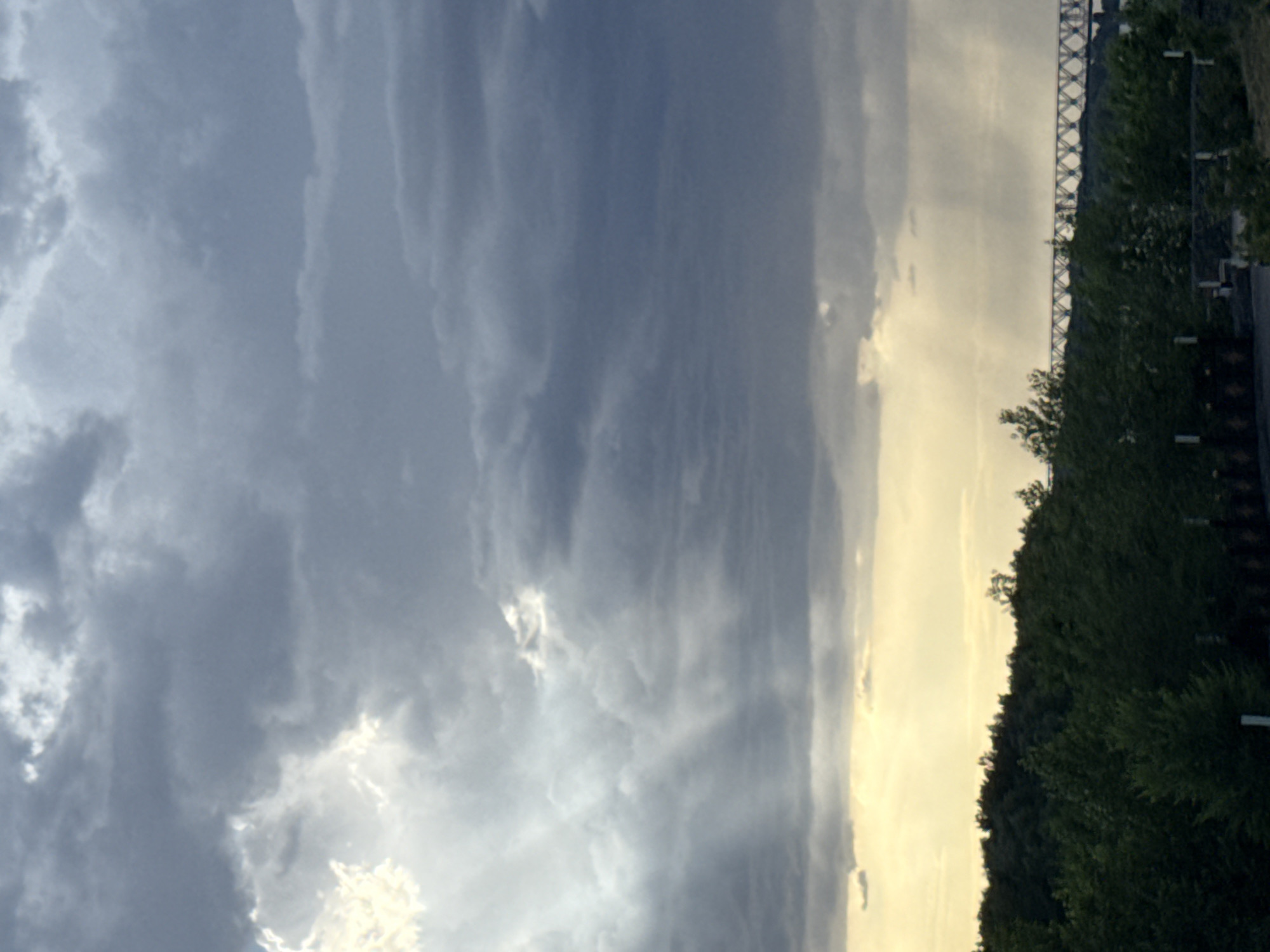 The image depicts a natural scene with a dramatic sky and a treeline. The sky is filled with a mix of dark, heavy clouds and lighter, more diffused clouds, suggesting an overcast or possibly stormy condition. Rays of sunlight break through in some areas, creating a luminous effect against the darker clouds. On the right side of the frame, a dense treeline forms a dark green boundary, with glimpses of what appears to be a road or pathway and some artificial lighting structures visible. In the distance, a section of a bridge or elevated structure with a lattice-like design is visible against the horizon, adding a man-made element to the otherwise natural landscape. The overall impression is one of a moody, atmospheric sky, possibly at dusk or dawn due to the interplay of light and shadow. The location context provided is Pavlodar, Kazakhstan. There is no visible text in the image.