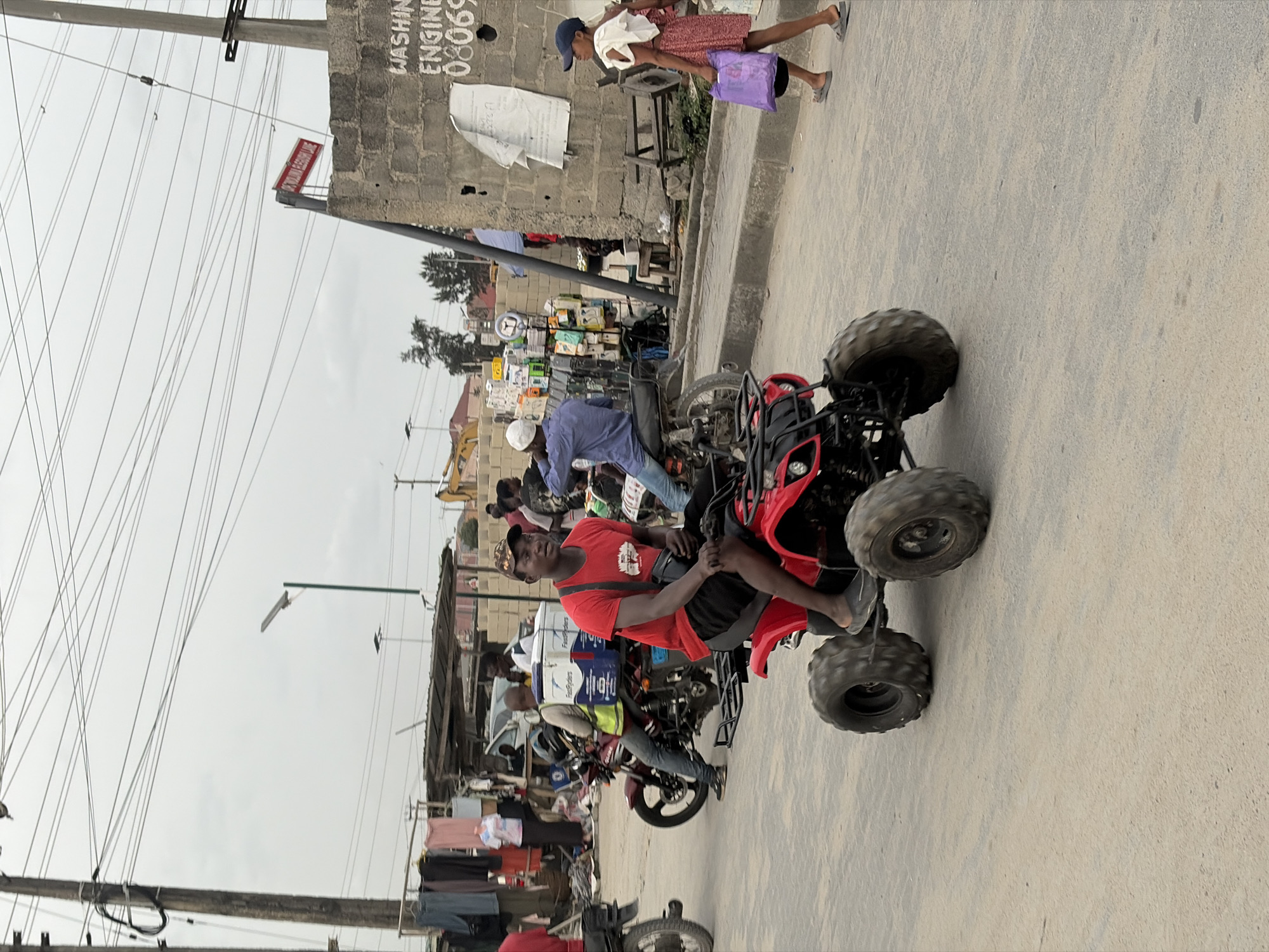 The image depicts a street scene in Igboefon, Nigeria. A man wearing a red shirt and a cap sits on a red ATV on a paved road. Behind him, there are several motorcycles, some with riders. A person wearing a white hat and a blue shirt is on one motorcycle. On the side of the road, there is a building with a sign that reads "Washing Engine" with a telephone number. A woman carrying a purple bag walks along the sidewalk near the building. Overhead, there are power lines and a street light. There is also clothing hung out to dry. The sky is overcast.