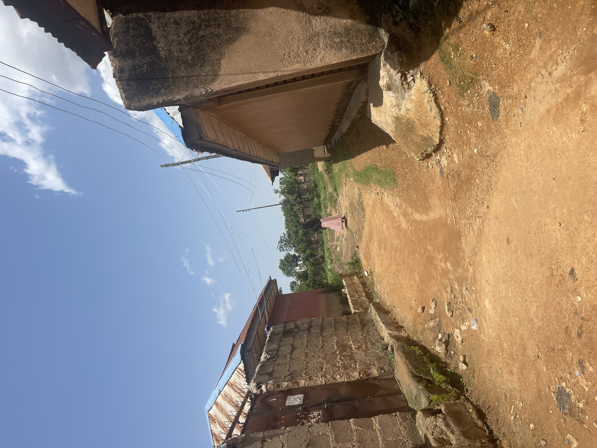 A person wearing a pink skirt and black head covering walks away from the camera down a dirt road between two buildings in Jos, Nigeria. The building on the left has a brick wall with a metal gate. The building on the right has a tan, stucco-like exterior. The sky is blue with scattered white clouds.