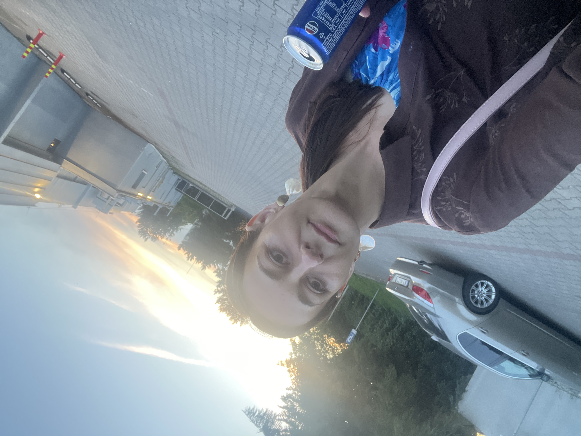 A woman is taking a selfie outdoors in what appears to be a parking lot. She is wearing a brown patterned shirt over a blue and pink floral dress. She has on large, statement earrings and is holding a can of Pepsi. To her left, a silver sedan is parked. The background shows trees and a building with a grey facade. The sky is a mix of blue and orange, indicating either sunrise or sunset. The ground is paved with interlocking grey bricks.
