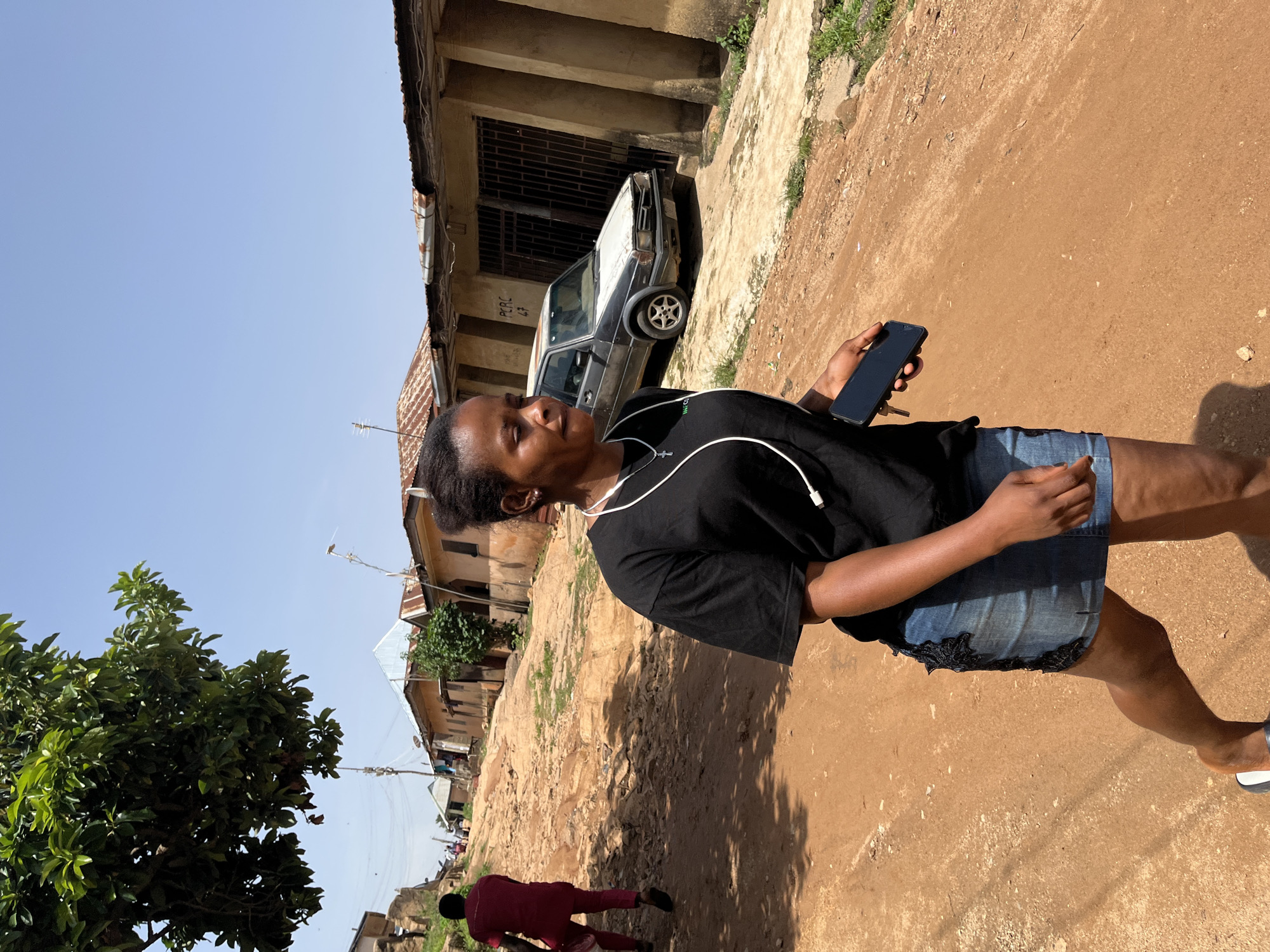 A young woman with dark skin and dark hair pulled back walks along a dirt road in Jos, Nigeria, on a sunny day. She is wearing a black t-shirt, a denim skirt, and white and black sandals. She has white earbuds in her ears and is holding a black smartphone in her right hand. Behind her, the dirt road slopes uphill, lined with modest buildings with corrugated metal roofs. A person in a red outfit is walking away from the camera up the road. Further up the road, a white and gray car is parked. The sky is clear and blue.