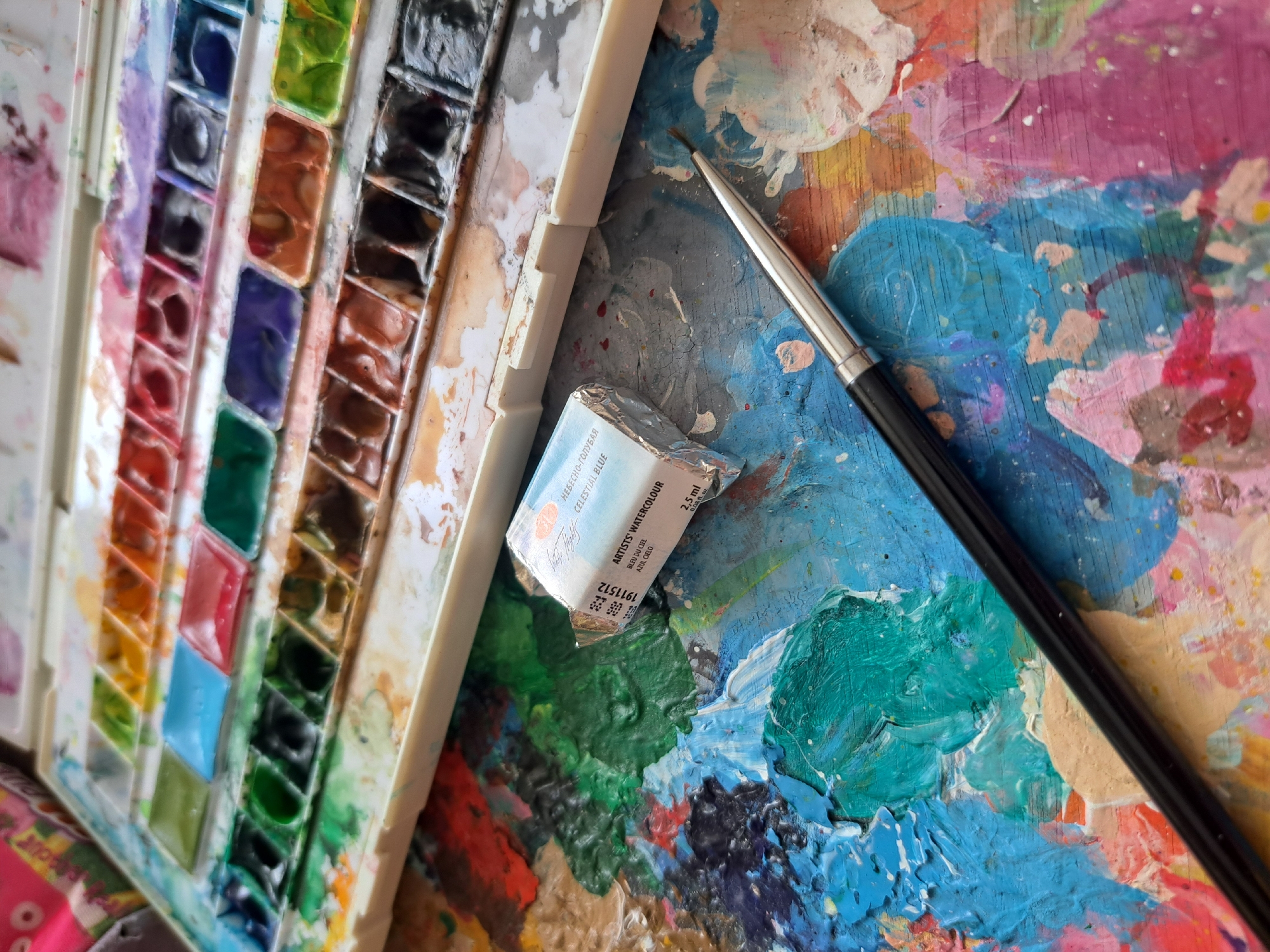The image displays a close-up view of art supplies, specifically a watercolor paint set and a palette loaded with colorful paints. In the foreground, a silver-handled paintbrush rests on a wooden palette covered in thick, textured strokes of various colors, including blues, greens, pinks, and yellows. A small, metallic-wrapped tube of watercolor paint is also visible on the palette, with visible text that reads "ARTISTS' WATERCOLOUR," "CELESTIAL BLUE," and "2,5 ml." To the left, a white plastic paint box is filled with individual pans of watercolor paints in a spectrum of colors, from vibrant reds and blues to earthy browns and blacks. The paint box appears used, with some colors showing signs of being frequently accessed. The overall scene suggests an artist actively engaged in painting, with the vibrant colors and textured palette conveying a sense of creativity and artistic process. There are no people visible in the image. The context of Popovo, Bulgaria, is external information not derivable from the image itself.
