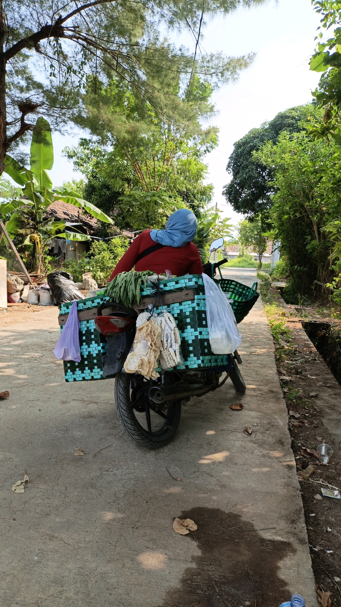 A person wearing a red long-sleeved shirt and a blue head covering is seated on a motorcycle. The motorcycle is equipped with a large green and blue woven basket attached to the rear, carrying produce and other items. Some items are hanging from the basket, including what appears to be dried fish or snacks and a purple plastic bag. In the background, there are trees, a house with a thatched roof, and a drainage ditch along a concrete path. The scene is lit by sunlight, casting shadows on the ground. The location is identified as Cikal, Indonesia.