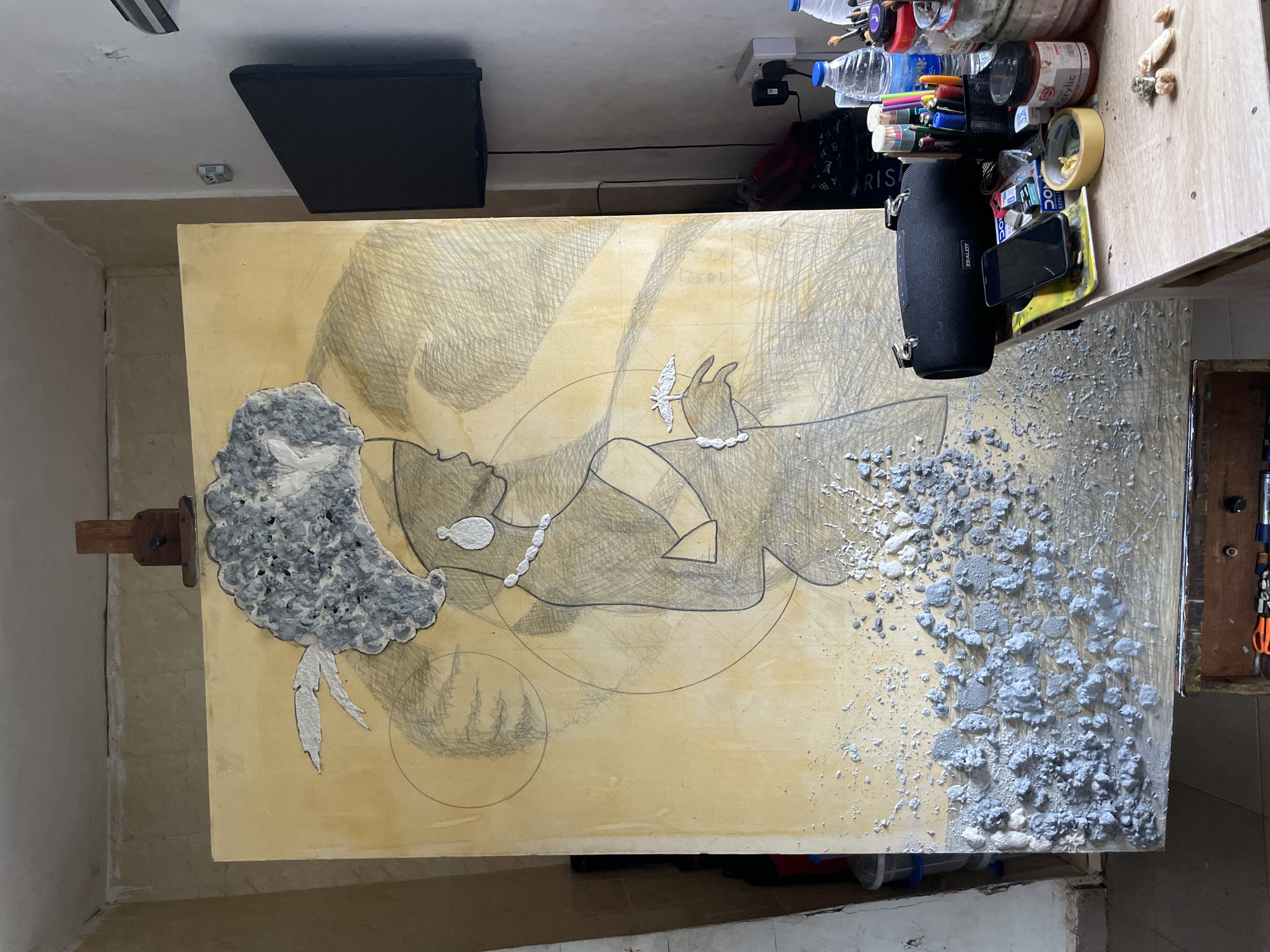 A large wooden board is oriented horizontally in a cluttered indoor space. The board bears a partially completed artwork depicting a reclining figure. The figure is outlined in pencil, with some areas filled in with grey paint or a similar textured material. The head of the figure, located on the left side of the board, has a prominent textured grey afro. The right side of the board is densely covered with the same grey textured material, giving the impression of it dissipating or flowing away from the figure.
Various objects surround the board, suggesting an artist's workspace. To the upper right, there are containers filled with art supplies, including pens, pencils, and bottles of paint. A black speaker rests on the board, and a smartphone is placed nearby. A black flat-screen monitor is mounted on the wall above the board.
The indoor space appears to be a studio or workroom. The walls are painted in neutral tones. The location is stated as Ado, Nigeria.
