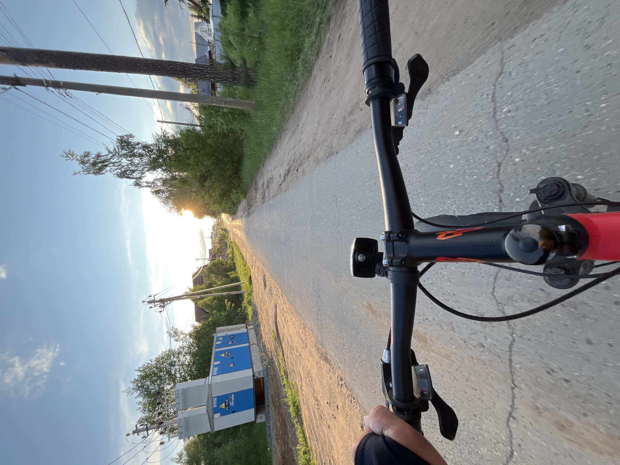 A first-person perspective image shows a bicycle moving along a straight road. The handlebars are visible, along with the rider's left hand. The road is flanked by vegetation on both sides, including grass, trees, and shrubs. Utility poles and power lines are on the left side of the road, along with a blue and white transformer. The sun is near the horizon, creating a bright spot in the distance along the road. The sky is a clear blue, with some wispy clouds near the horizon. Based on known information and the image context, the location is Chaikovsky, Russia.