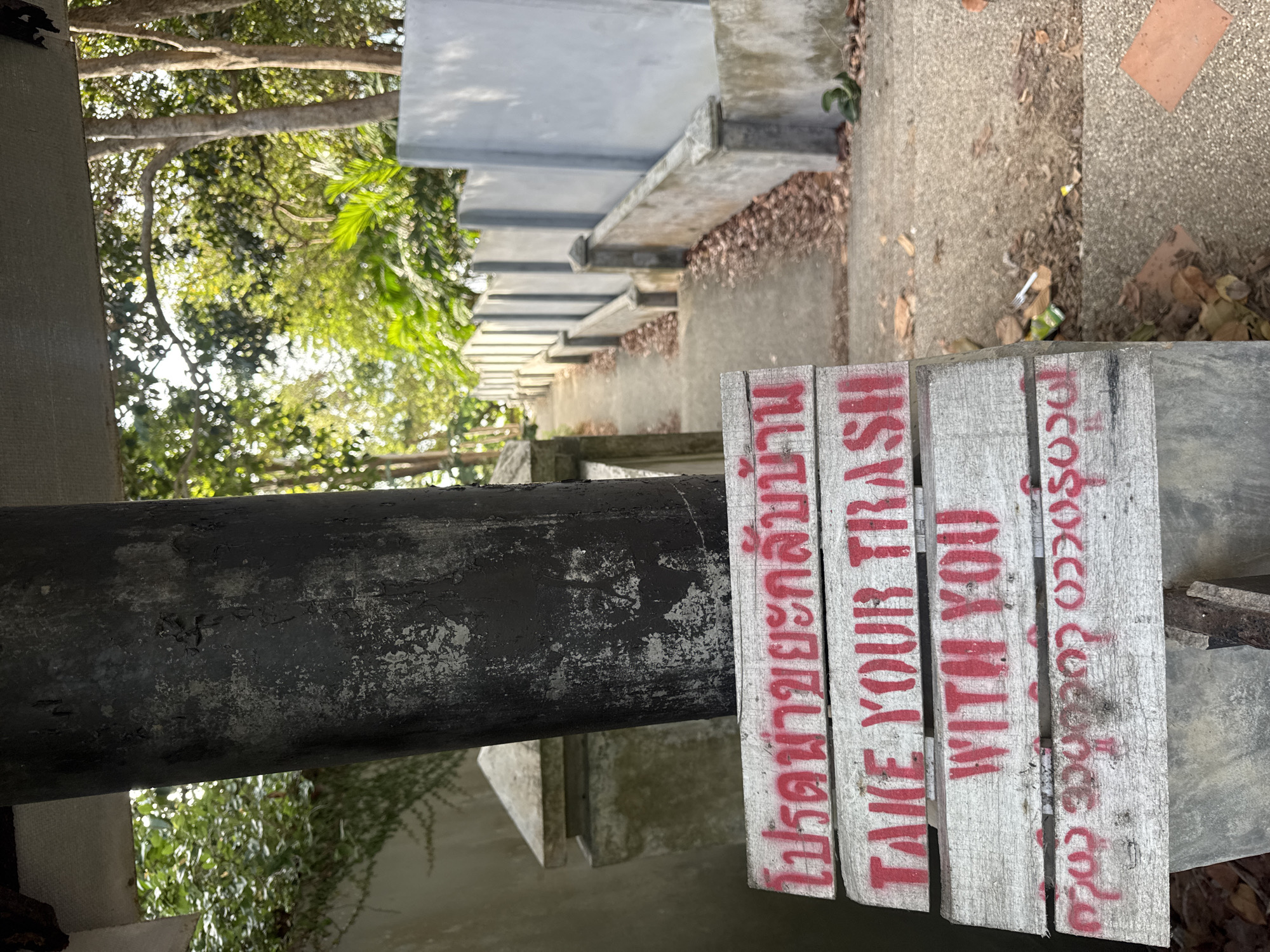 A weathered, white wooden sign with red lettering is positioned in the foreground, leaning against a dark, cylindrical structure. The sign displays text in three languages. The top line, in Thai script, reads "โปรดนำขยะกลับบ้าน" (Please take your trash home). The middle line, in English, states "TAKE YOUR TRASH WITH YOU". The bottom line appears to be in Burmese script. Behind the sign, a concrete walkway ascends a slope, flanked by a low concrete wall and a taller, gray concrete wall with vertical segments. The path is covered with fallen leaves. Lush green foliage and trees are visible in the background, suggesting an outdoor environment with a tropical or subtropical climate. The overall scene depicts an outdoor pathway with a clear directive to dispose of waste responsibly.