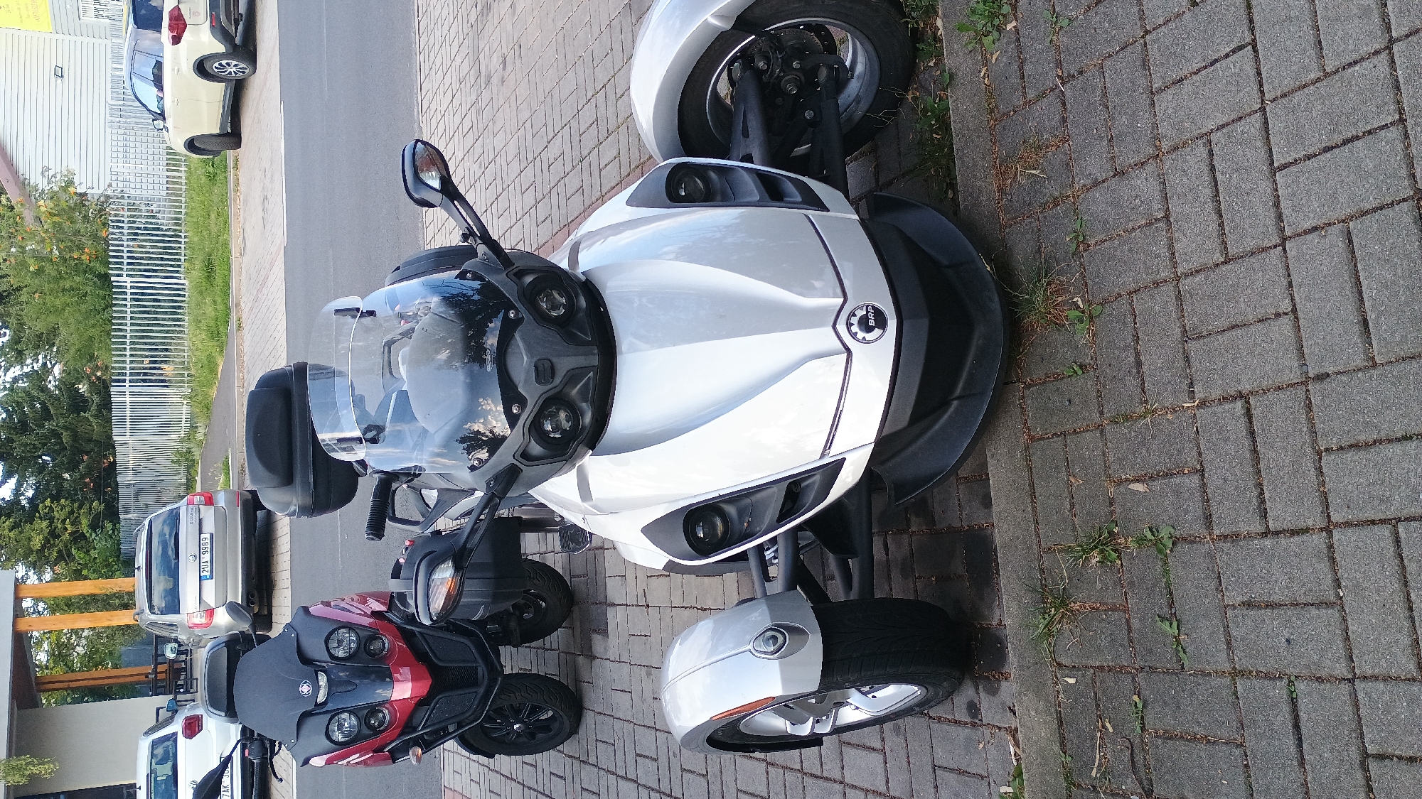 In Sokolov, Czechia, two three-wheeled scooters and two cars are parked on a paved area next to a street. The scooter in the foreground is white with a black windshield and front fairing, featuring a "BRP" logo. To its left is a red and black scooter with a black windscreen and storage box. Behind the red and black scooter is a silver Skoda Octavia sedan with license plate "2UA 9989". Further in the background, a cream-colored Suzuki Vitara SUV is parked. The scene appears to be a public area, possibly a parking lot, with a white fence and trees visible in the background.