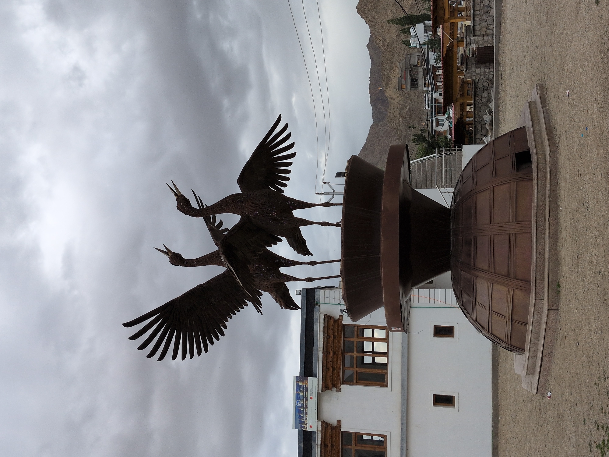 In the city of Leh, India, a large metal sculpture of two cranes with their wings spread stands prominently in an outdoor courtyard. The sculpture is dark brown and appears to be made of intricately woven wire or metal strips. The cranes are positioned on a tiered, dark brown pedestal, which in turn rests on a domed, metallic base with a grid-like pattern.
To the left of the sculpture is a white building with traditional wooden window frames and ornate carvings above them. A sign with text and images is affixed to the side of this building. The ground in front of the sculpture and building is a sandy, earthy tone.
In the background, a range of brown, mountainous terrain rises. Several buildings, including some with modern architectural elements and others with a more traditional style, are visible in the middle ground, nestled at the base of the mountains. Power lines stretch across the sky. The sky itself is overcast with a layer of grey clouds. The scene appears to be devoid of any visible people or animals, other than the metallic cranes.