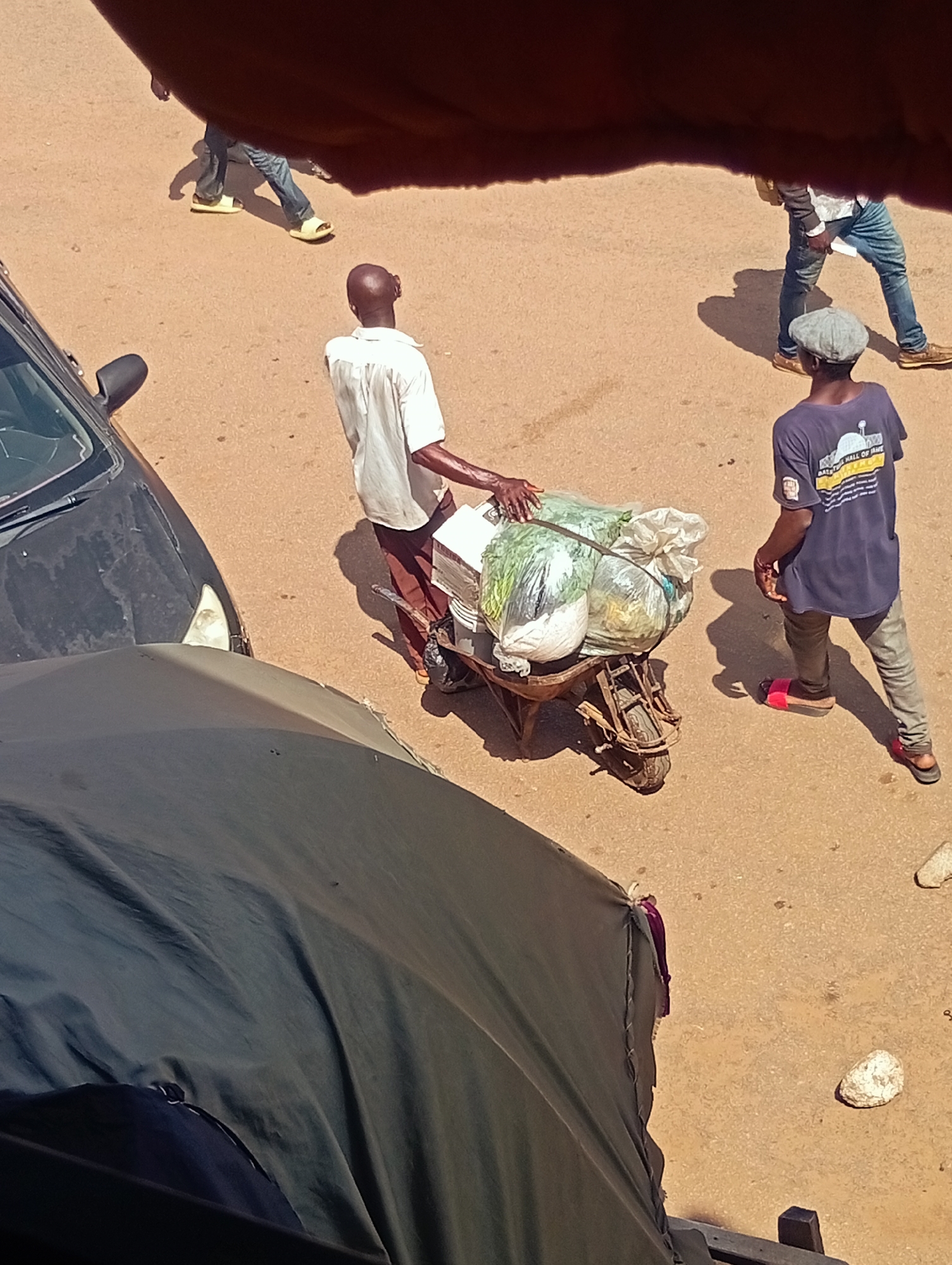 In Bukuru, Nigeria, a man in a white shirt and brown pants pushes a wheelbarrow filled with produce. The wheelbarrow has one wheel and is loaded with what appears to be vegetables wrapped in plastic bags. Another man, wearing a dark blue t-shirt and gray pants, stands to the right of the wheelbarrow, facing away from the viewer. He wears a flat cap and red sandals. In the background, another person in jeans and sandals walks away from the scene, casting a shadow on the sandy ground. A black car is partially visible on the left side of the image, and a large dark green tarp covers the bottom portion of the frame, obscuring the ground and some of the objects. The overall scene depicts a marketplace or street environment with people engaged in commerce or transportation of goods.