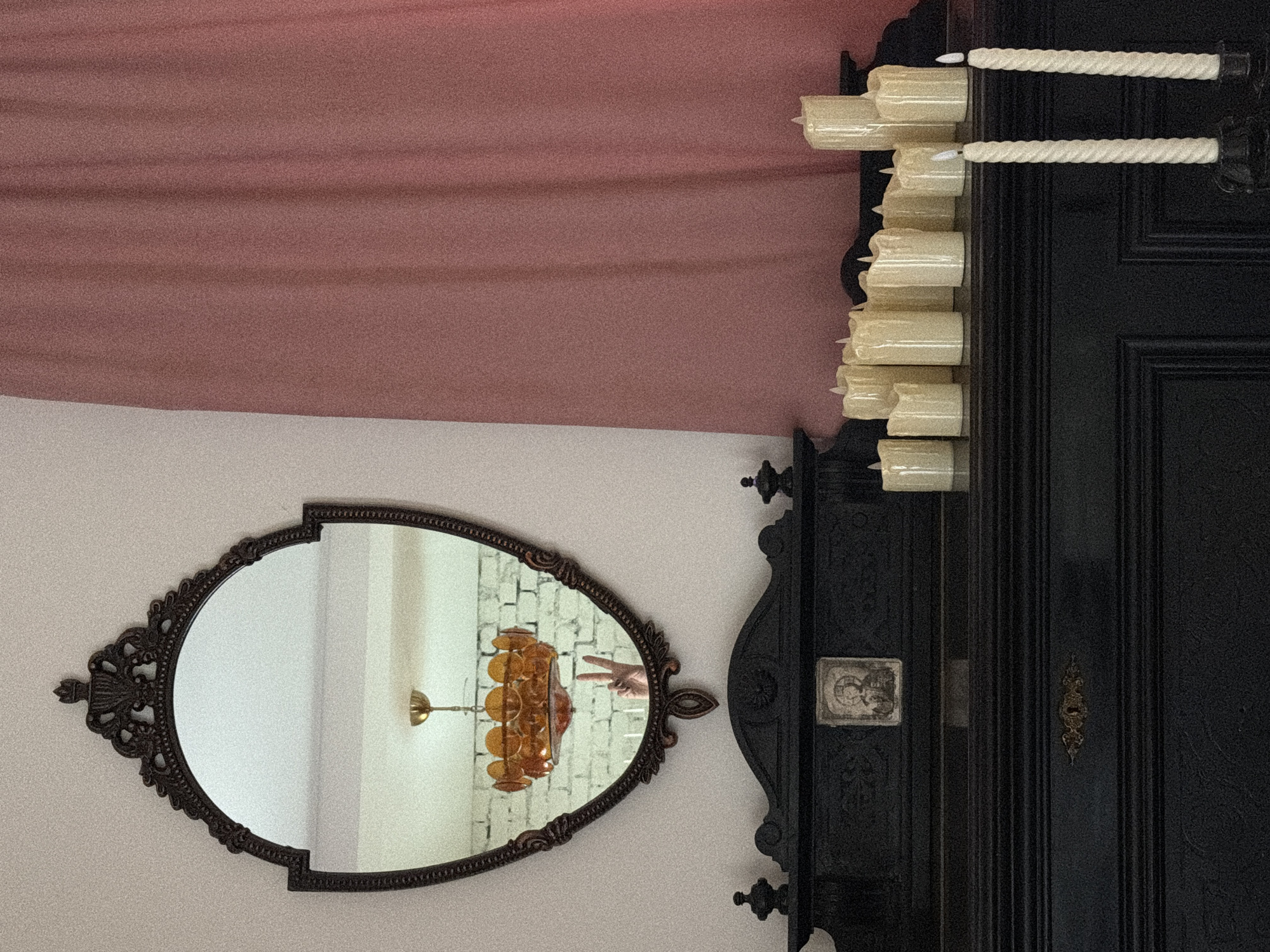 The image depicts an indoor scene, likely a room in a home in Kolomna, Russia. In the center, a large, ornate dark wood mirror hangs above a dark wood mantelpiece. The mirror reflects a vintage amber chandelier and a hand making a peace sign against a white brick wall. On the mantelpiece, a small framed religious icon is placed centrally. To the right of the icon, and extending across the width of the mantelpiece, are numerous ivory-colored pillar candles of various heights. Two tall, twisted ivory candles in dark candleholders are positioned in the foreground, to the right. A dusty rose-colored curtain hangs on the right side of the image, partially obscuring the wall. The lighting is soft, with the primary illumination appearing to come from above and the right. The overall atmosphere is one of a dimly lit, possibly vintage or gothic-themed interior.
