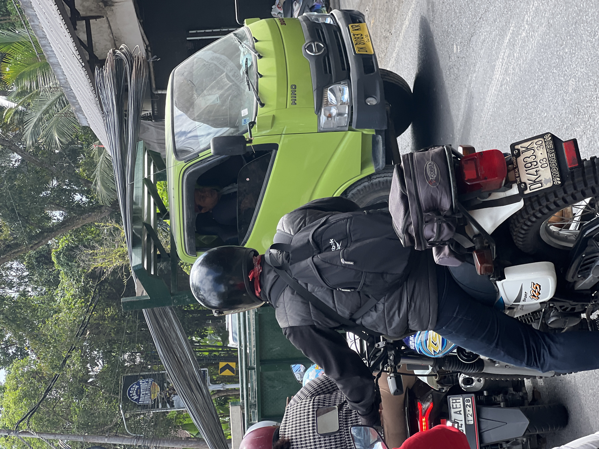 A bright green Hino truck carrying a load of metal bars is stopped on a street in Payangan, Indonesia. The truck's license plate is DK 8983 KR. In front of the truck, two motorcycles are also stopped in traffic. The rider of the first motorcycle, seen from behind, wears a black helmet, a black vest over a black long-sleeved shirt, and blue jeans. A black backpack is on their back. The second motorcycle is partially visible, with its rider wearing a patterned shirt and a red helmet. The surrounding environment shows dense green foliage and trees, indicating a tropical setting. The street appears to be paved, and power lines are visible overhead. A sign for "MASON ADVENTURES" is visible on the left side of the image. The overall scene depicts a typical traffic situation on a street in Payangan.