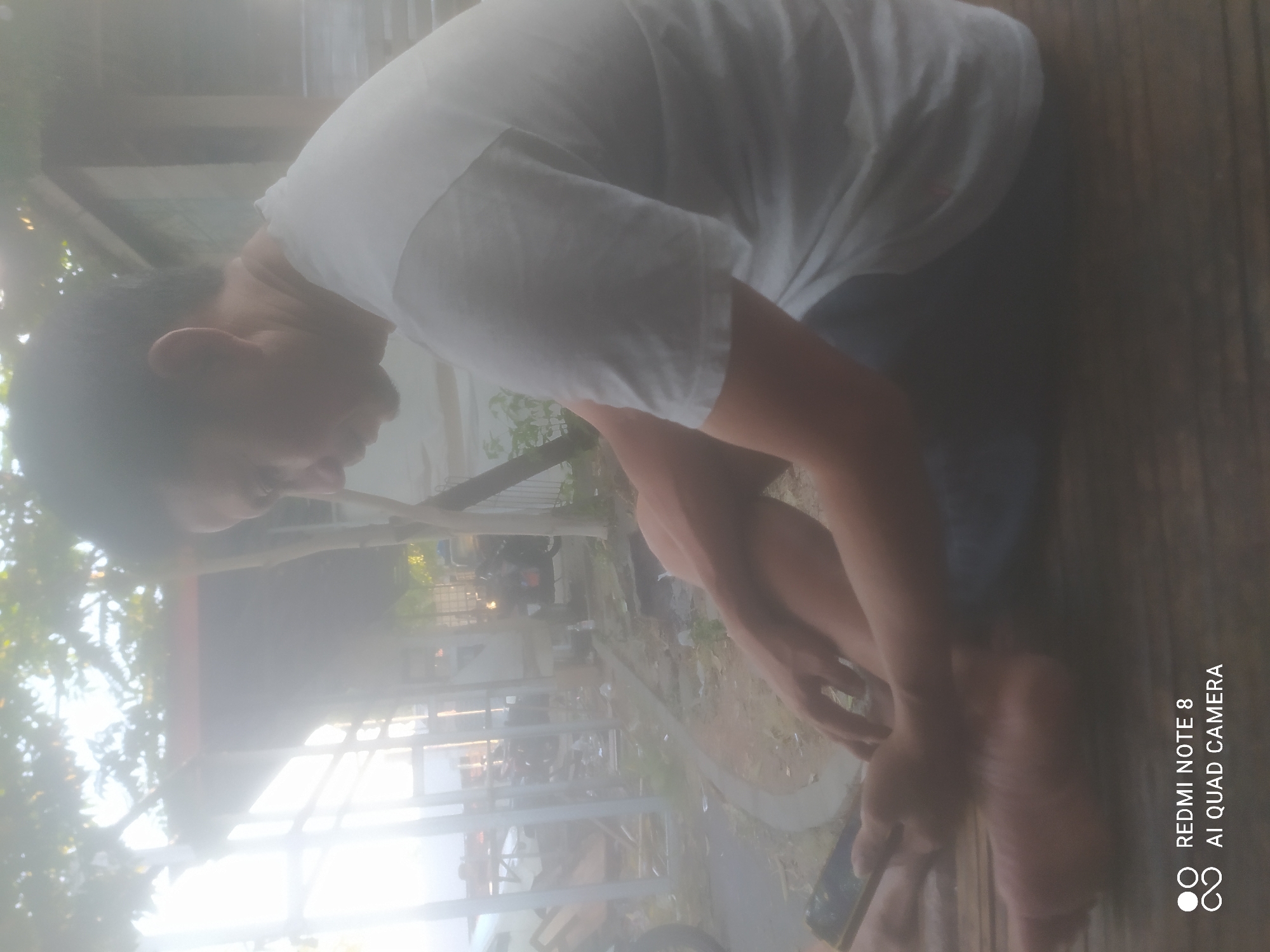 A person is sitting on a wooden surface, their legs crossed. They are wearing a white t-shirt and blue pants. The person is holding a smartphone in their left hand and appears to be looking down at it. The background shows an outdoor environment with trees, structures, and what appear to be motorcycles or scooters. The image has a hazy or slightly overexposed quality. The text "REDMI NOTE 8 AI QUAD CAMERA" is visible in the bottom left corner, indicating the device used to capture the photograph. The scene appears to be in Sumurber, Indonesia, as indicated by the provided context.