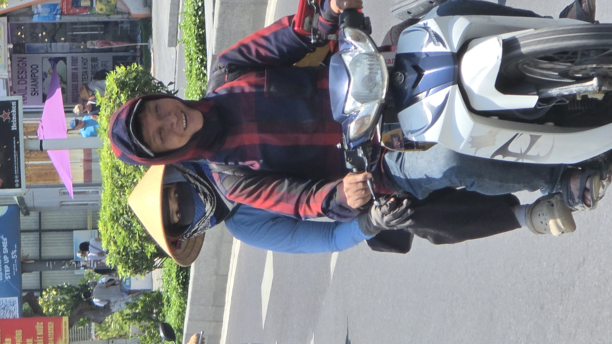 The image shows two people on a motorcycle in Nha Trang, Vietnam. One person, wearing a traditional conical hat and face covering, is driving. The other person, wearing a hooded jacket, is riding as a passenger and smiling. The motorcycle is blue and white. In the background, there are buildings with advertisements and signs, including those for Heineken, "TAIL DESIGN SHAMPOO", and "MASSAGE BODY & FOOT". Other text in Vietnamese and possibly Russian can also be seen. The side of the street has green shrubbery. The motorcycle is on a grey asphalt road. Text "PHONG 01/4/2026" and "STEP UP SMES" are partially visible on signs.