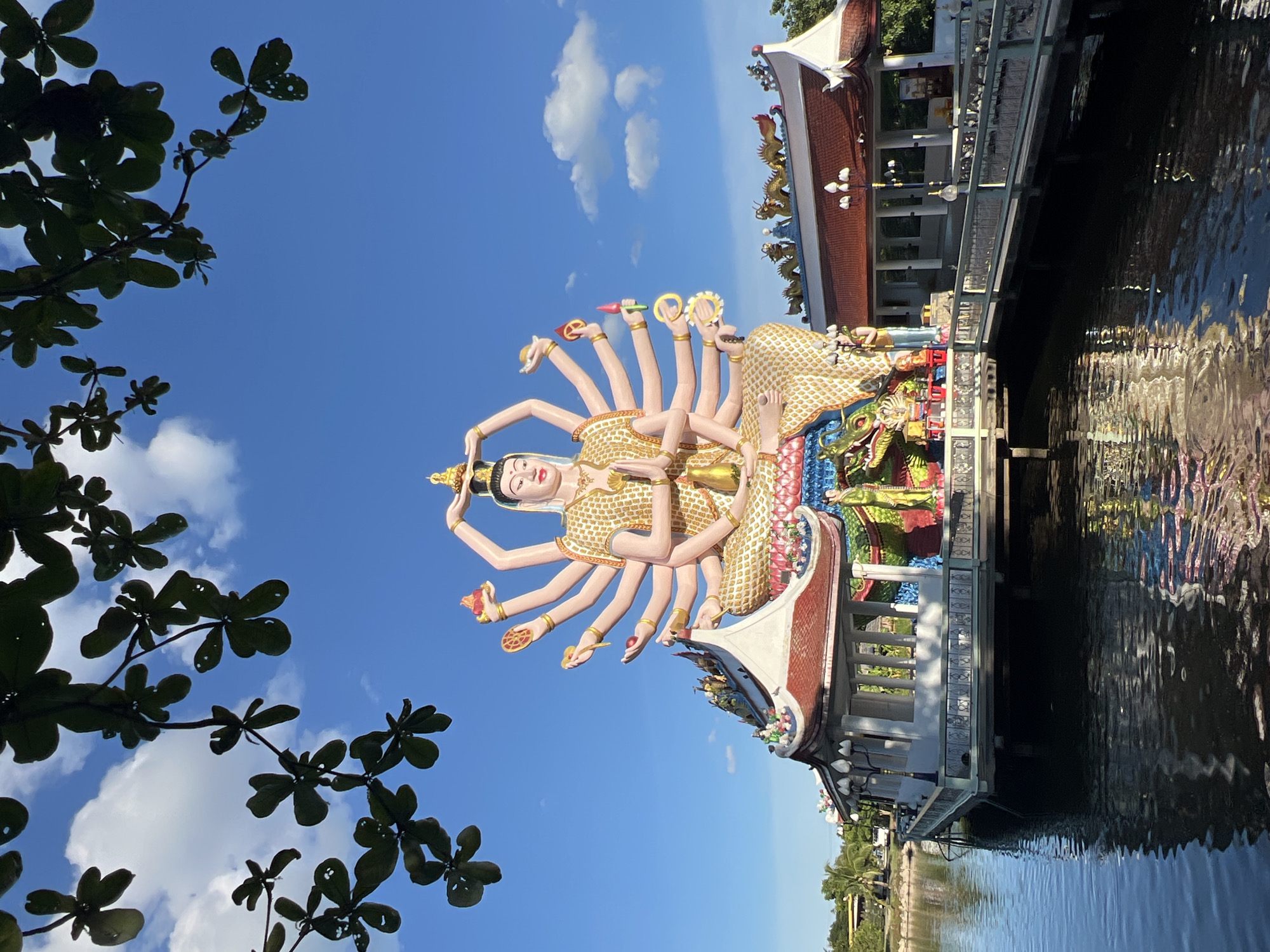 This vertical, outdoor, eye-level medium shot captures a vibrant statue with multiple arms, possibly depicting a deity, against a clear blue sky. The statue is adorned in a textured gold and white garment and is positioned in front of a building with traditional Thai architecture, featuring red tiled roofs and decorative dragon sculptures. A body of water with rippling reflections is visible to the right of the building. Silhouetted leaves from a tree in the foreground frame the top left portion of the image. The bright sky and sunlight suggest it is daytime, likely with good weather. The overall impression is one of spiritual or cultural significance in a tropical setting. There is no visible text in the image.