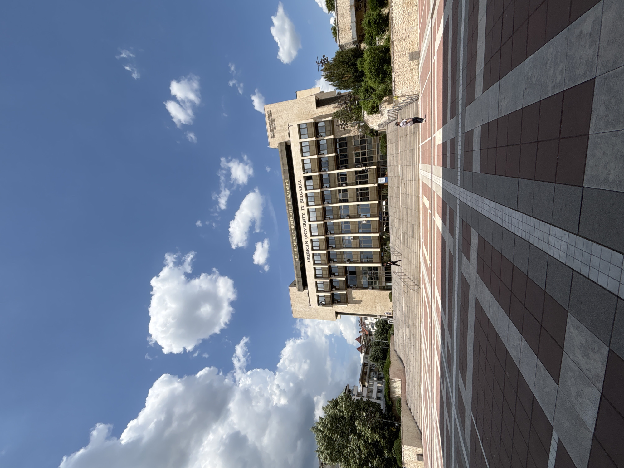 The image shows the American University in Bulgaria (AUBG) campus. The building is a beige stone structure with multiple floors of windows and a green sign. A large, patterned plaza is in the foreground. The sky is blue with scattered white clouds. One person is walking down a set of stairs towards the building. Other buildings and trees are in the background.