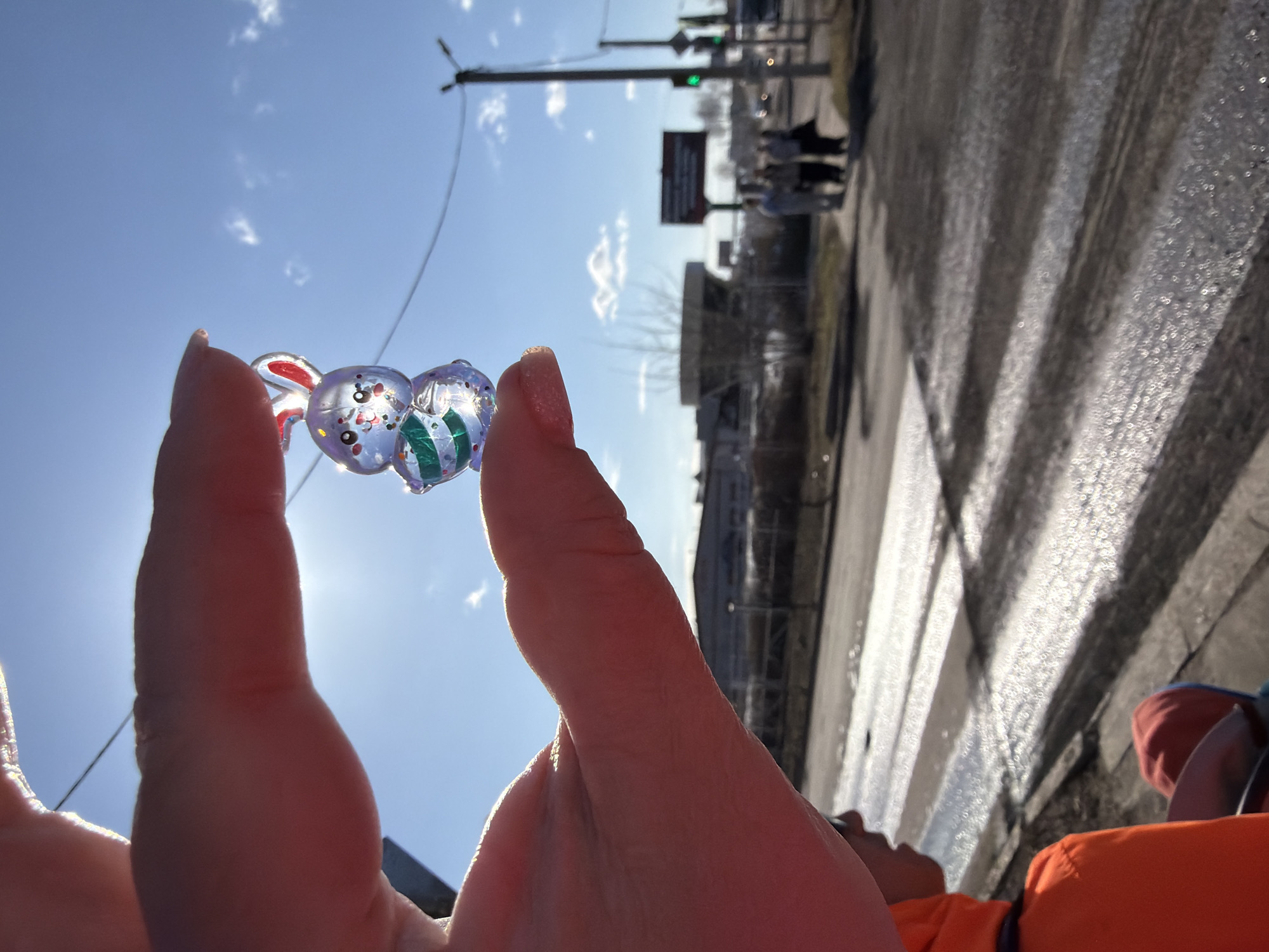 The image is taken at ground level in Chaikovsky, Russia, likely near a pedestrian crossing. In the foreground, a hand holds a small, transparent toy featuring two cartoonish figures, one shaped like a bunny with red ears and the other with green stripes. Sunlight shines through the toy, creating glare. In the background, the scene is out of focus, but a street can be discerned with visible pedestrian crosswalk markings, buildings, traffic lights, and a sign. Two people wearing white clothing can be seen crossing the street. The road surface appears wet, possibly from rain or melting snow. In the bottom of the frame, an orange piece of clothing is visible. The sky is light blue with sparse clouds.