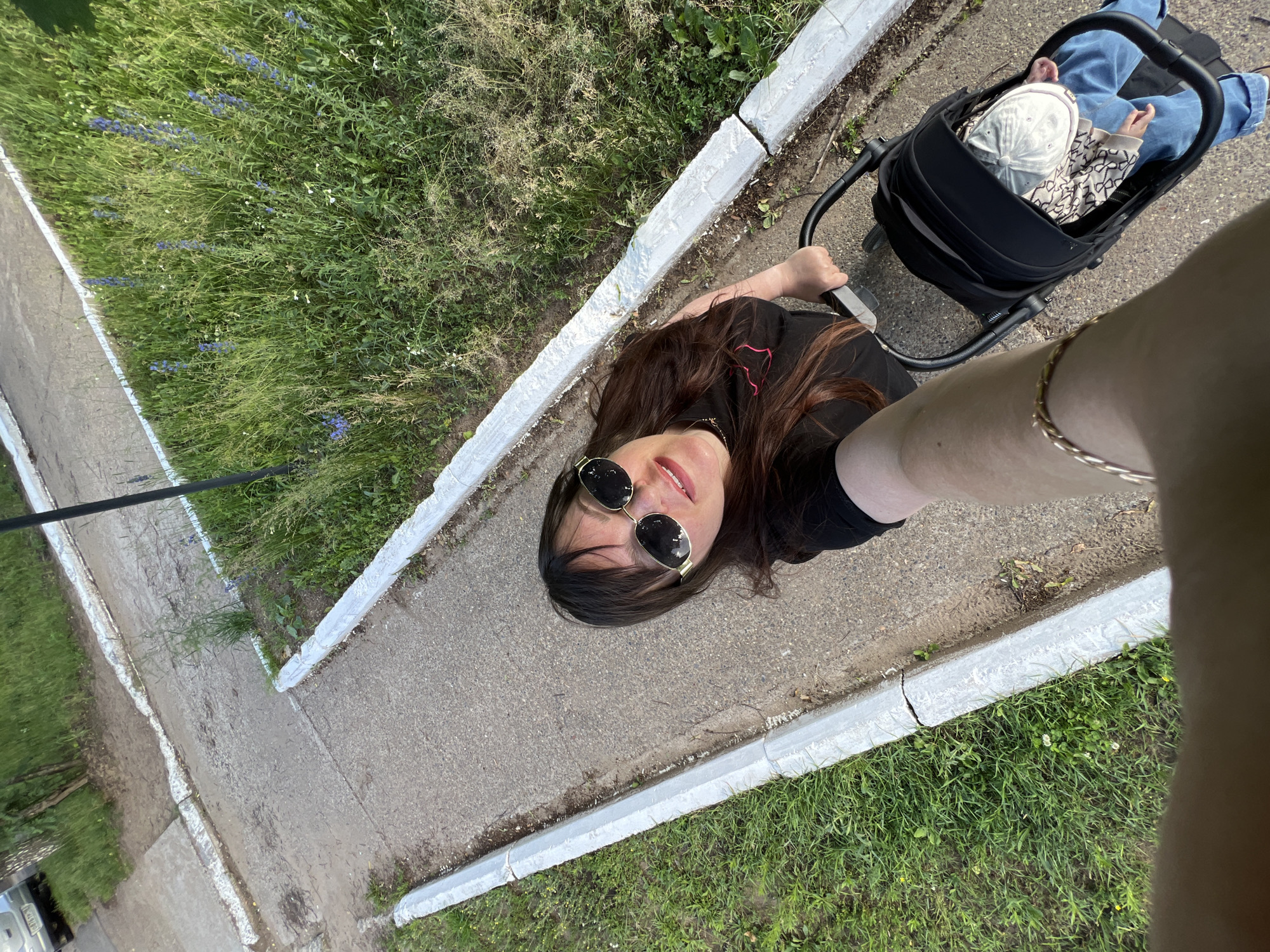 In this overhead shot, a woman is pushing a stroller with a baby inside, walking along a paved path bordered by grass and white curbs. The woman, wearing sunglasses and a black t-shirt, is looking at the camera, smiling. The baby, wearing a light-colored cap and blue jeans, is nestled in the stroller. The scene appears to be outdoors, possibly in a park or residential area, with lush green grass and some small blue flowers visible. The pavement is grey and textured, and the white curbs show signs of wear. The overall lighting suggests it is daytime, likely with clear weather. There is no visible text in the image.