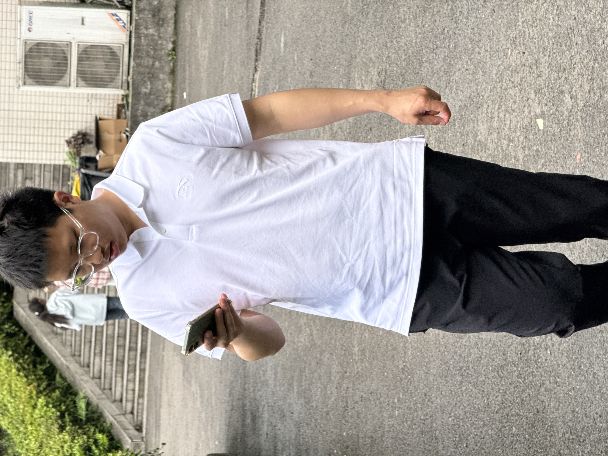 A young man stands outdoors, holding a smartphone. He is wearing glasses, a white polo shirt with a small logo, and black pants. He is positioned on an asphalt or concrete surface. In the background, a staircase is visible, along with some greenery and a building with air conditioning units. Another person is walking on the staircase in the background. The location may be Jiangjin, China, based on the background scenery.