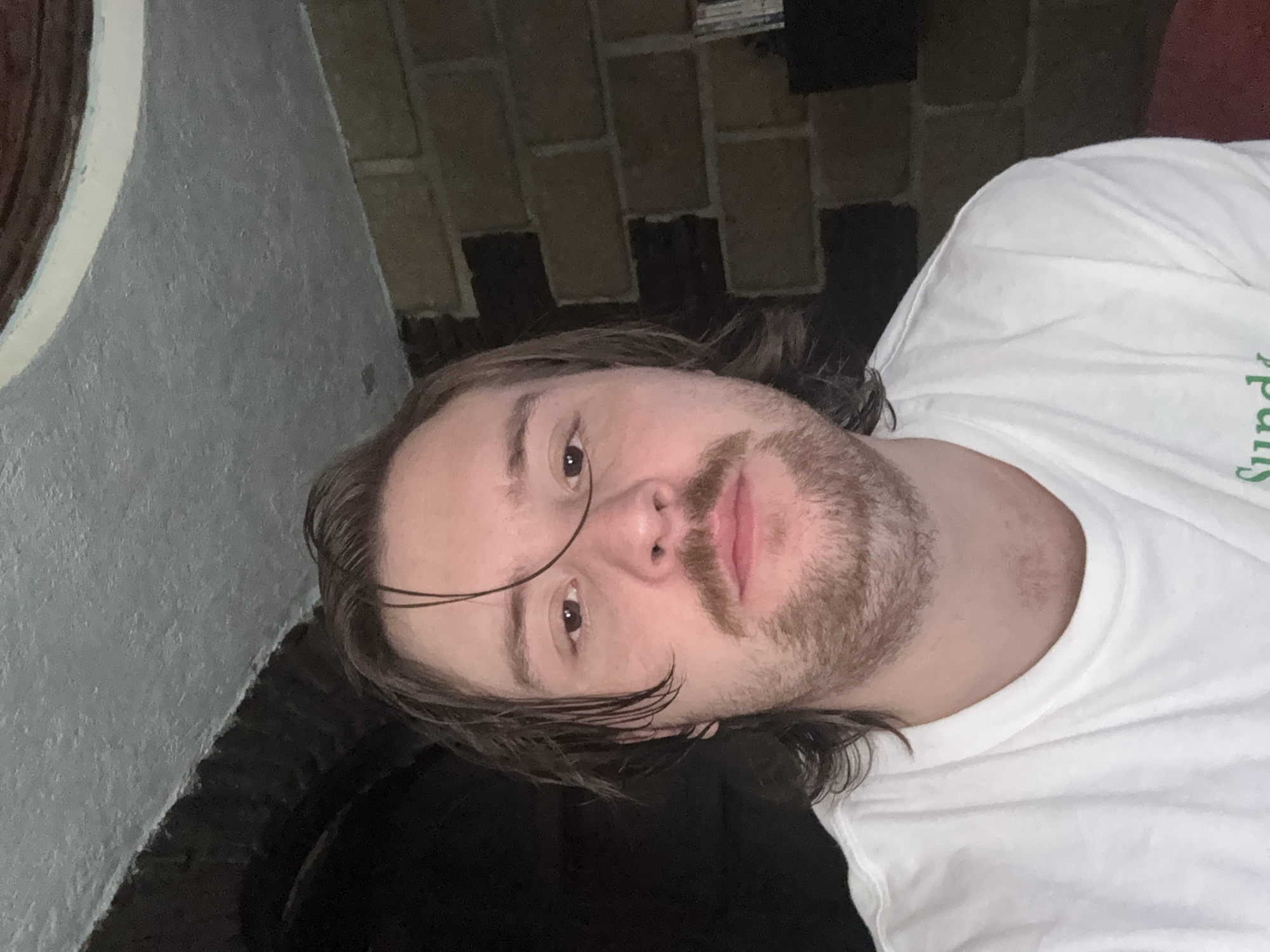 A person is shown in a selfie. The individual is wearing a white t-shirt and has brown hair, a mustache, and a beard. The background includes a brick wall and a section of what appears to be a concrete structure. The location in the city of Cañada de Madero, Mexico, is unknown.