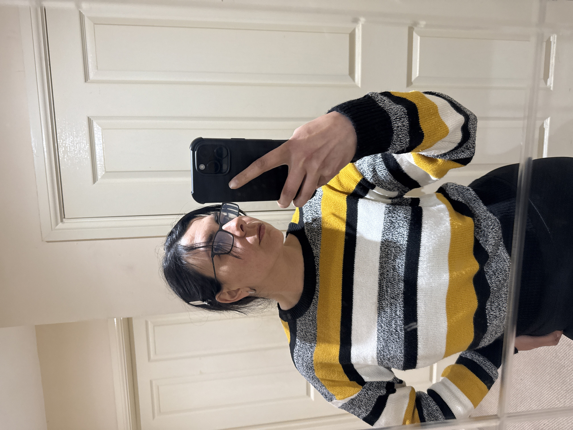 A woman with dark hair and glasses is taking a mirror selfie with a black phone in a room with two white paneled doors. She is wearing a striped sweater with yellow, white, and black bands. The location in the city of Great Yarmouth, United Kingdom is unknown from the image.
