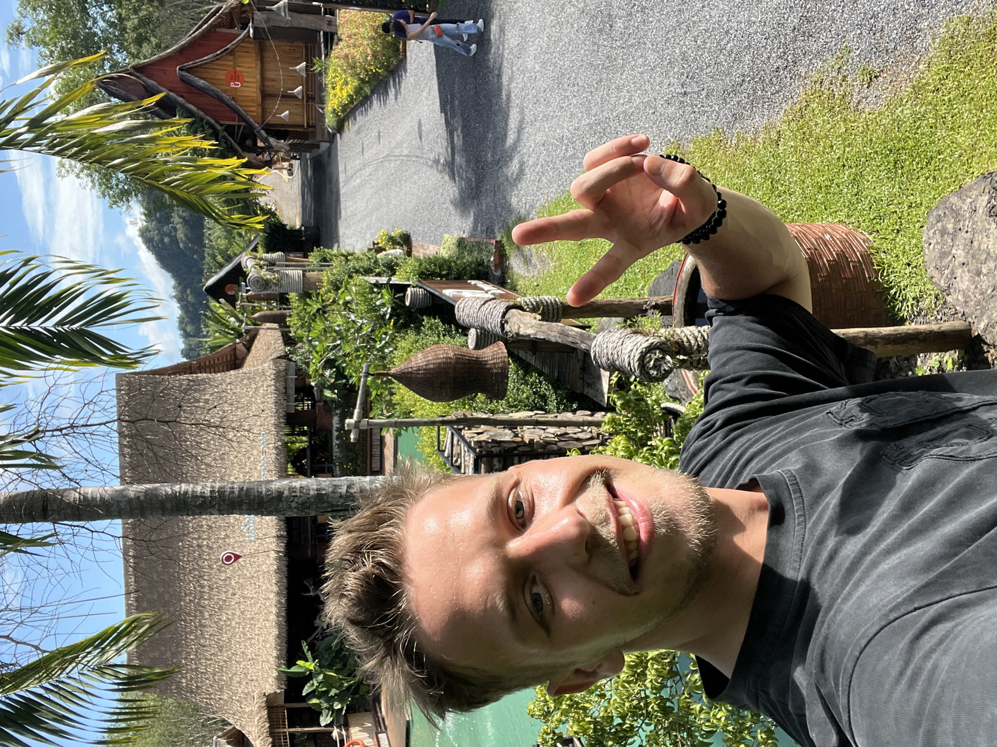 In Tambon Pa Kae Bo Hin, Thailand, a young man with blond hair and a mustache smiles at the camera, making a peace sign with his right hand. He wears a black t-shirt and a black beaded bracelet. In the background, a turquoise swimming pool is visible, surrounded by lush green foliage. Several thatched-roof buildings with wooden accents are scattered throughout the landscape, some featuring decorative elements. A gravel path winds through the scene, with two individuals walking in the distance. The sky is clear and blue with some clouds, and mountains rise in the background.