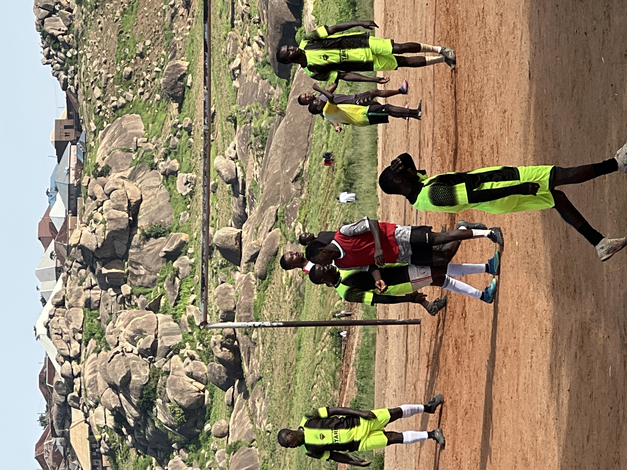 In Jos, Nigeria, a group of young men are playing soccer on a dusty field. The field is located at the base of a rocky hill, with houses and trees visible in the background. The sky is clear and the sun is shining. The players are wearing bright, colorful jerseys, and their movements suggest an active game. The scene captures a moment of recreation and camaraderie in an outdoor urban setting.