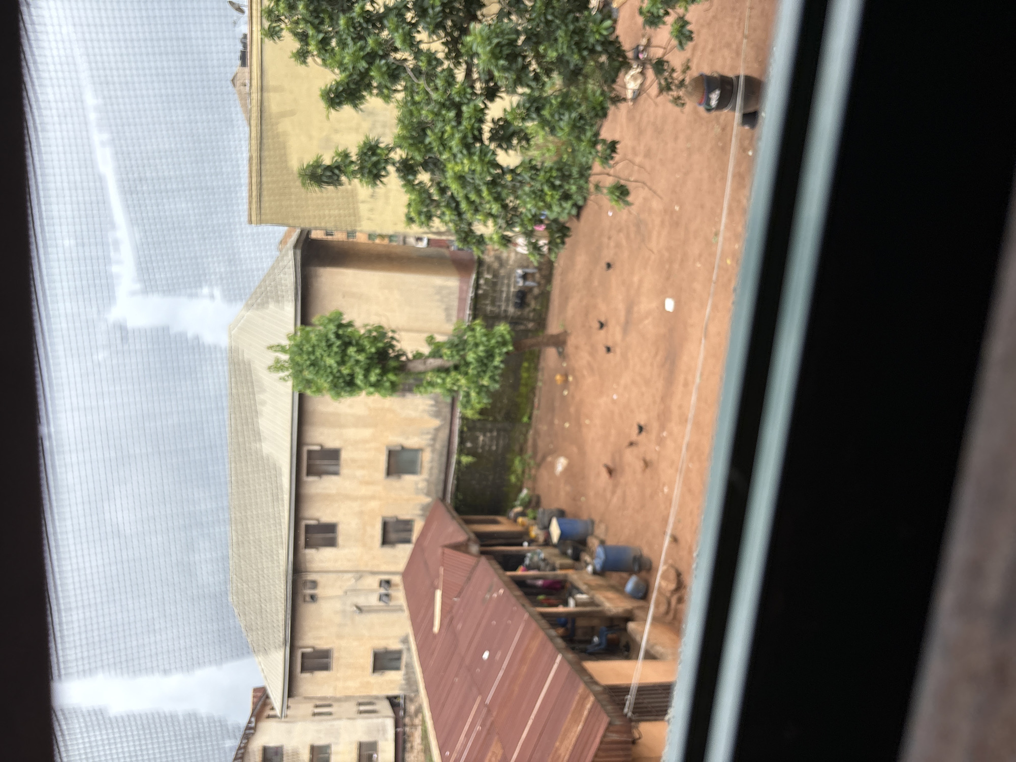 The image depicts a courtyard scene in a residential area of Onitsha, Nigeria, viewed through a window with a screen. In the background, two buildings are visible. The one on the left is a light brown, multi-story structure with several windows. Adjacent to it, and slightly closer, is a larger, yellowish building, also with windows. A tall, slender tree stands in front of the yellowish building, and to the right, a dense green tree with many branches obscures part of the building.
In the foreground of the courtyard, a single-story structure with a corrugated red metal roof extends from the left side of the image. This structure appears to be an open-air market or workshop, with several people visible inside and under its overhang. Blue barrels and other containers are placed around the area. The ground is covered in reddish-brown earth, and several chickens are scattered across it. A thin wire runs horizontally across the courtyard. On the far right, a dark object, possibly a person wearing dark clothing and a hat, is partially visible behind a tree. The sky above is overcast with white clouds.