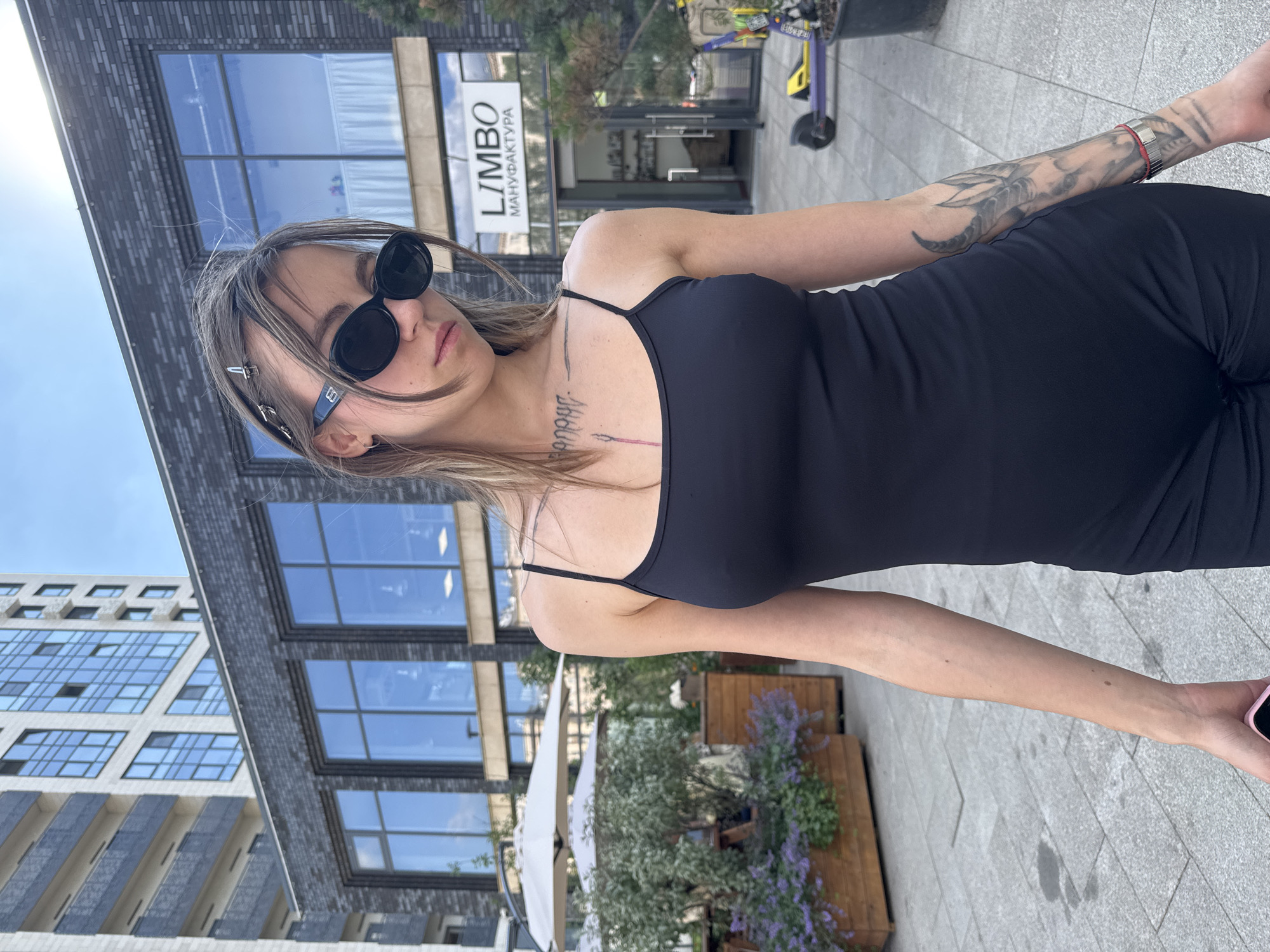 In Moscow, Russia, a young woman with long, light brown hair, wearing sunglasses and a black tank dress, stands on a paved walkway. She has tattoos on her chest and right arm. Behind her, a dark brick building with large windows is visible, with signage that reads "LIMBO MANUFACTURA". To the left of the building, there are outdoor seating areas with umbrellas and planters filled with purple flowers and greenery. In the background, a tall, modern apartment building with balconies and blue-tinted windows can be seen. A scooter is parked near the building. The sky is overcast.
