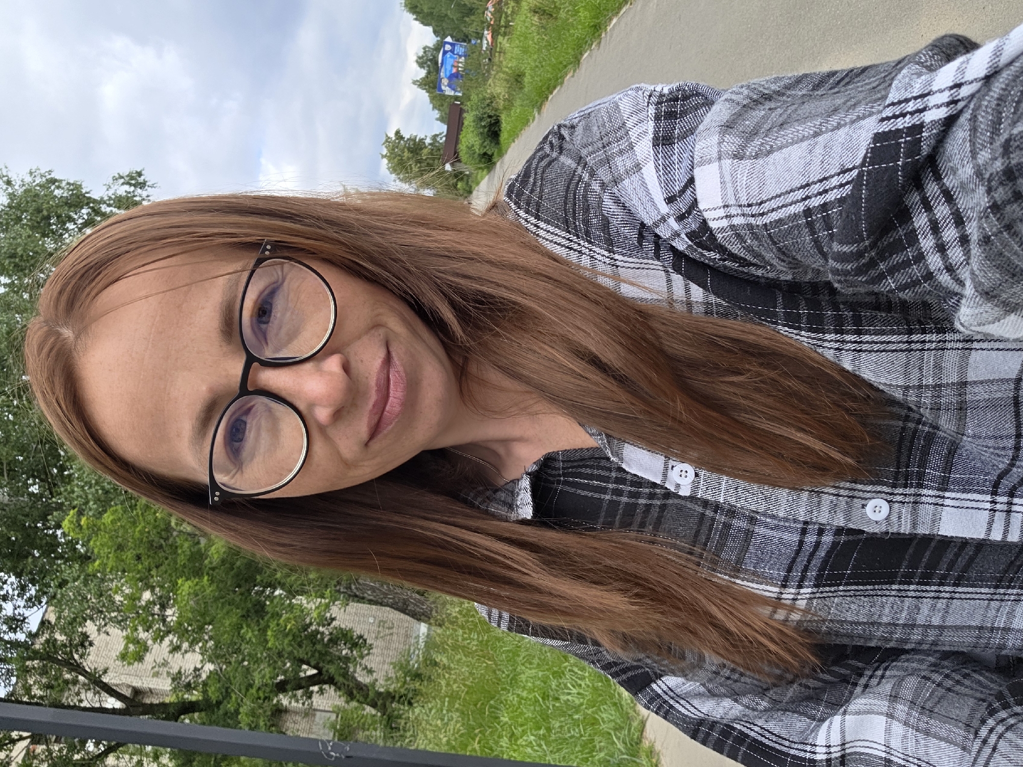 A medium shot captured outdoors shows a woman with auburn hair wearing a black and white plaid shirt and glasses. She is standing on a paved path, with a grassy area and trees to her left, and a road, more greenery, and buildings in the background to her right. The sky is overcast. The image is likely taken in the city of Chaikovsky, Russia, based on the surrounding environment.