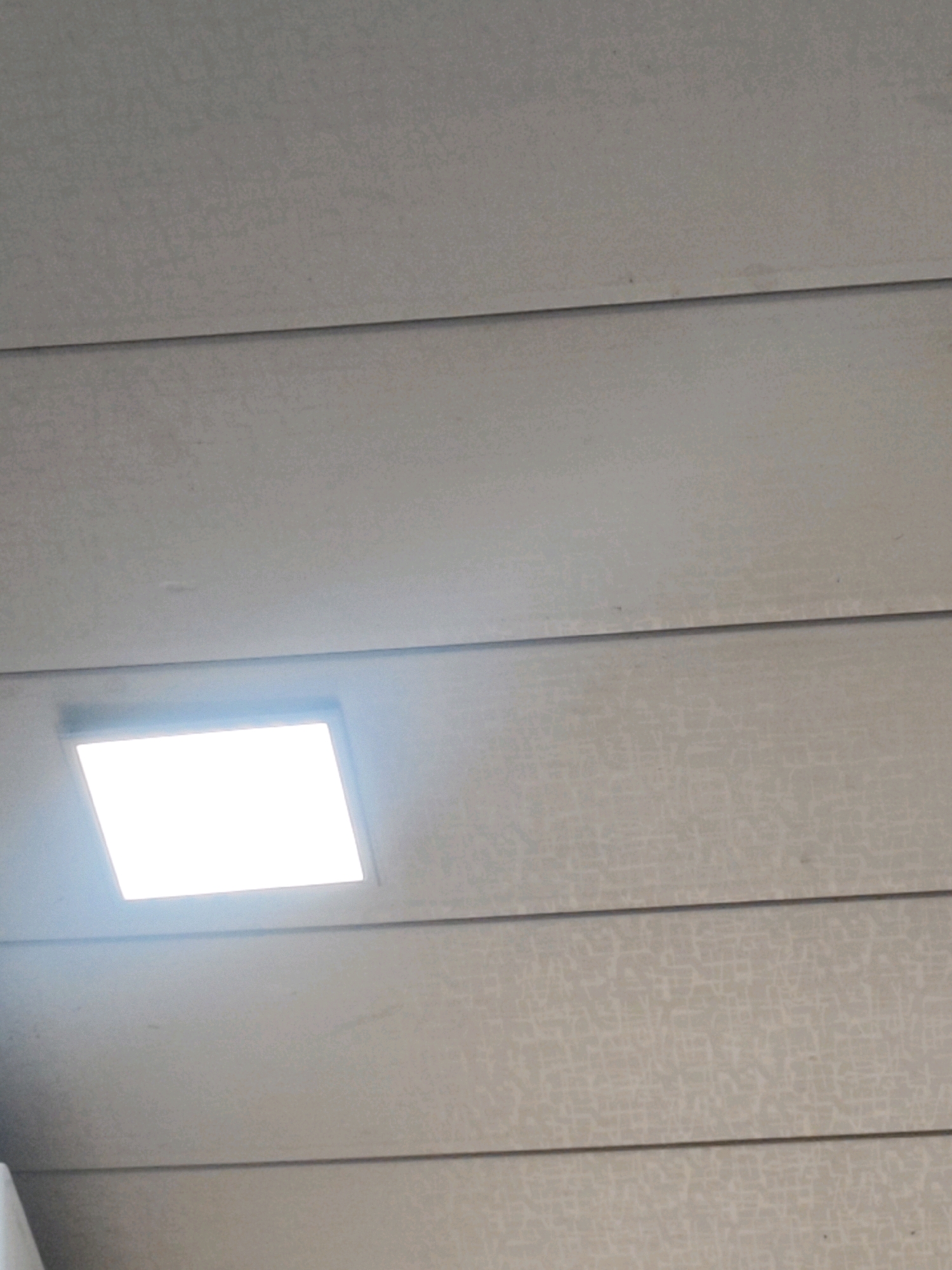 The image shows a close-up view of a plain, light-colored ceiling with horizontal paneling. A bright, rectangular light fixture is embedded in the ceiling in the lower left quadrant of the image. The lighting is diffuse and appears to be coming from an overhead source. The texture of the ceiling is visible, with subtle patterns or graining on the panels. There are no people or any other objects visible in the image. The environment appears to be indoors and possibly a modern building due to the recessed lighting. No specific location within Sumerpur, India, can be determined from the image.