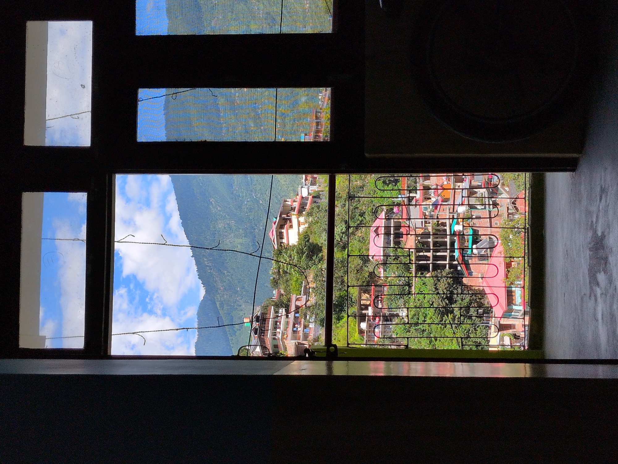 An outdoor scene of Kullu, India is captured through a window. The view is of a hillside covered with houses and lush green trees, with a backdrop of majestic mountains under a clear blue sky with scattered clouds. The houses are built in a traditional Indian architectural style, with multiple stories, balconies, and red-tiled roofs. A metal railing runs along the edge of the balcony, in front of the window. The window itself is divided into multiple panes, some of which are covered with netting, suggesting the presence of mosquitoes. The foreground is dark, with the window frame and part of a wall visible, suggesting the photo was taken from inside a building. The overall impression is one of a serene and picturesque mountain town, with a blend of natural beauty and human habitation.