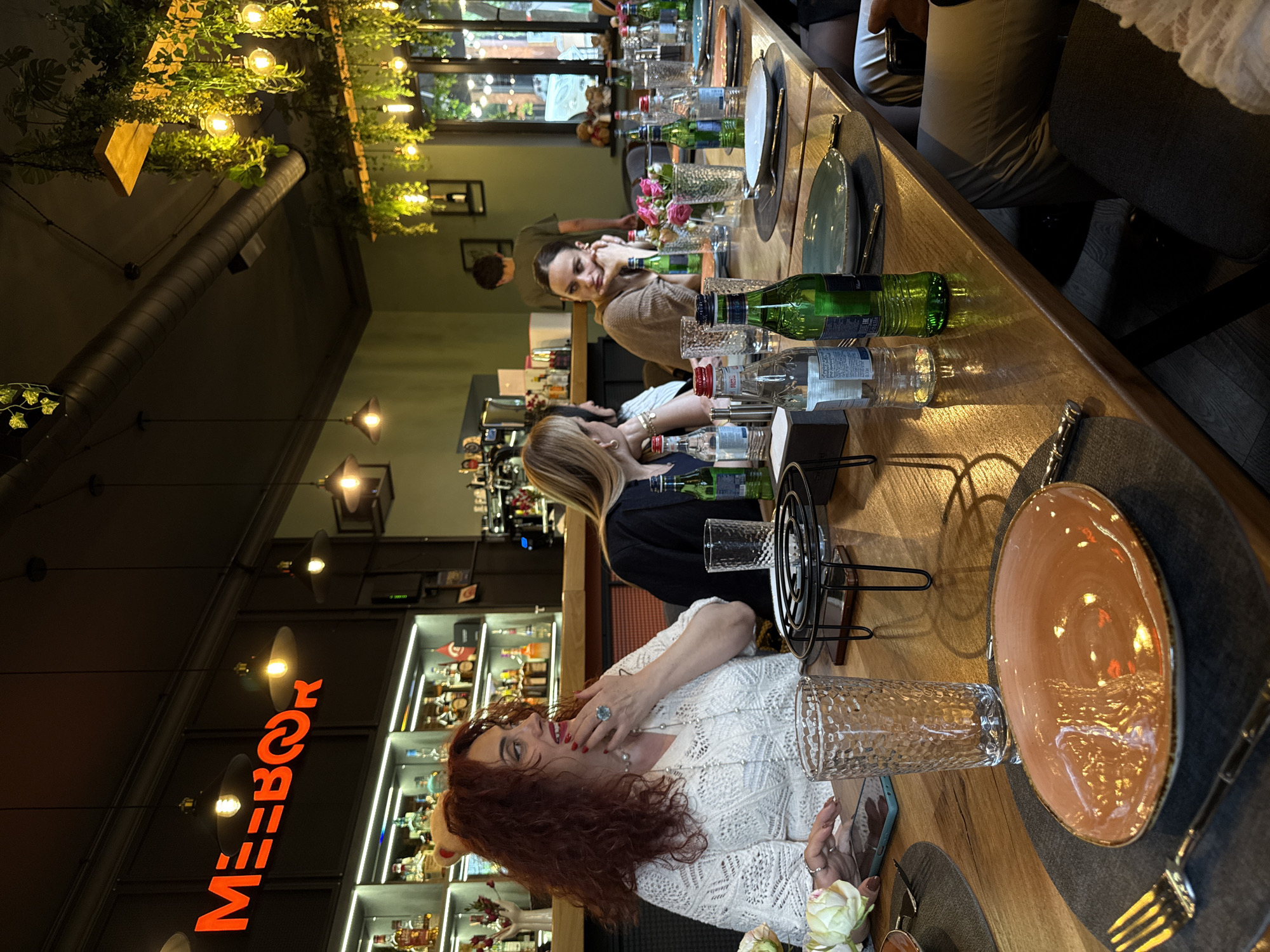 The image shows a group of people at a restaurant or bar, likely in Yerevan, Armenia, based on the reversed "MEZZO" sign visible. A group of women are seated at a long wooden table set with plates, glasses, water bottles, and decorations, suggesting a dinner or celebration. The lighting is a mix of natural and artificial, with decorative lights and greenery hanging from the ceiling. The background features a well-stocked bar with various bottles and decorative items.