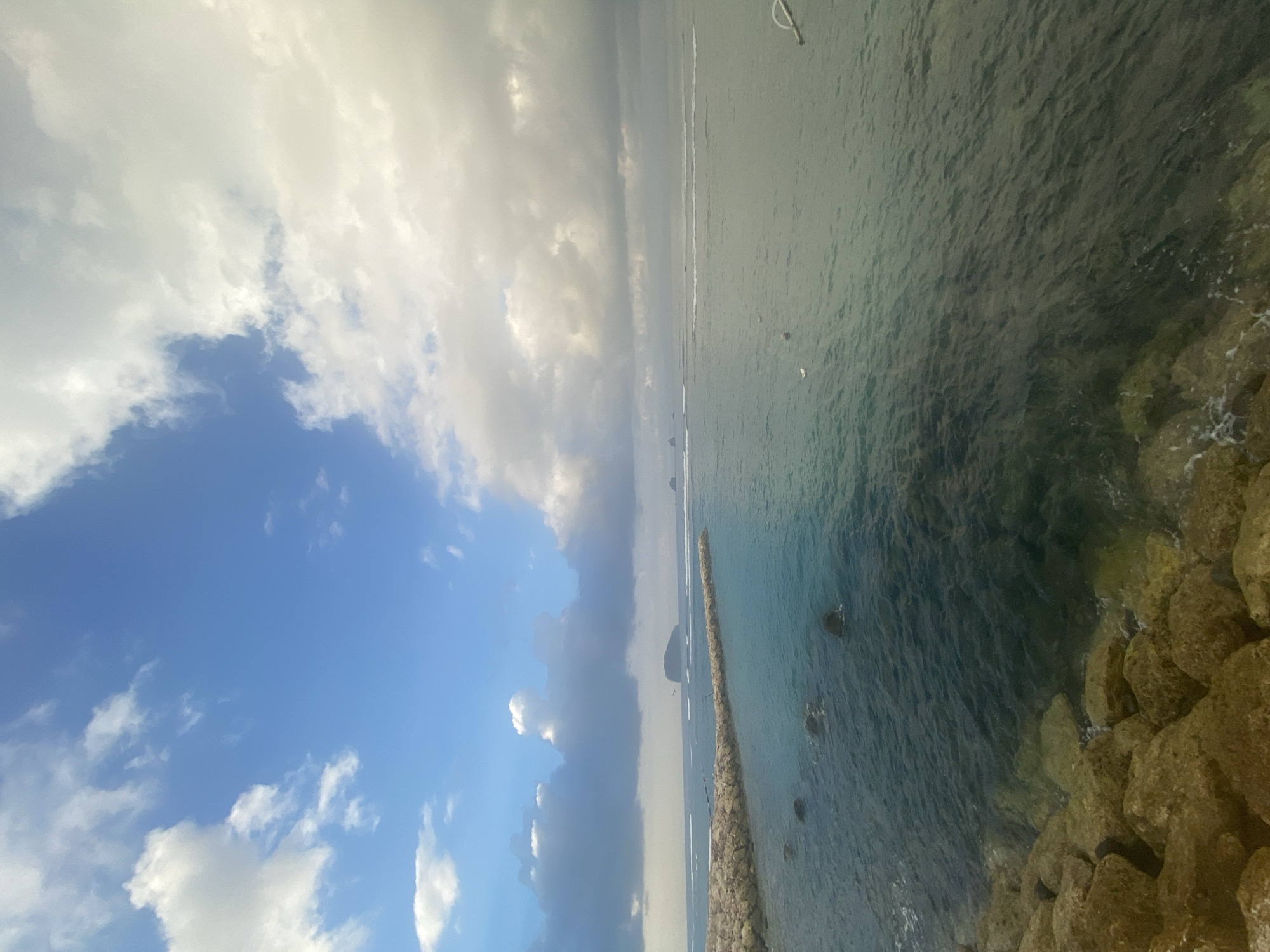 The image depicts a coastal scene with a breakwater made of large rocks extending into the ocean. The water is clear and turquoise near the shore, gradually becoming darker blue further out. In the distance, several small islands are visible on the horizon, shrouded in a light mist. The sky is a mix of blue sky and scattered white and gray clouds, with some darker clouds suggesting overcast conditions. The foreground is dominated by the rocky shore. This scene is likely located in Karangasem, Indonesia, known for its beautiful coastline and proximity to islands.