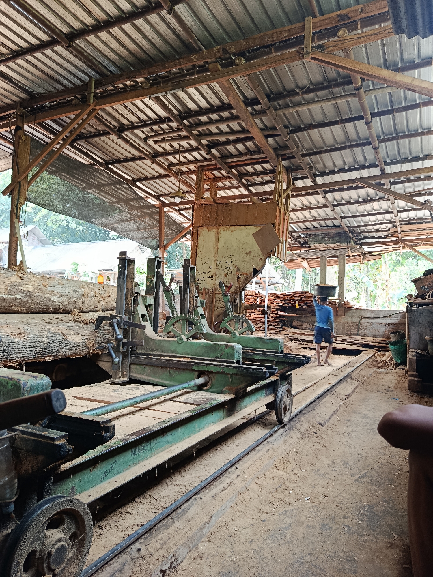 In the city of Kasihan, Indonesia, this image captures the interior of a lumber mill. The scene is dominated by a large, industrial sawmill machine, painted green, with a complex system of tracks and pulleys. The machine is positioned on a dirt floor covered in sawdust.
To the left of the sawmill, several large logs are stacked, awaiting processing. Behind the machine, stacks of processed lumber are visible, indicating ongoing operations.
In the background, a person wearing a blue shirt and dark shorts carries a large, circular object on their head, walking towards the right side of the frame. They appear to be engaged in moving materials within the mill. The environment is partially covered by a corrugated metal roof supported by wooden beams, suggesting an open-air or semi-enclosed structure. The overall impression is one of an active, rustic industrial setting focused on woodworking.