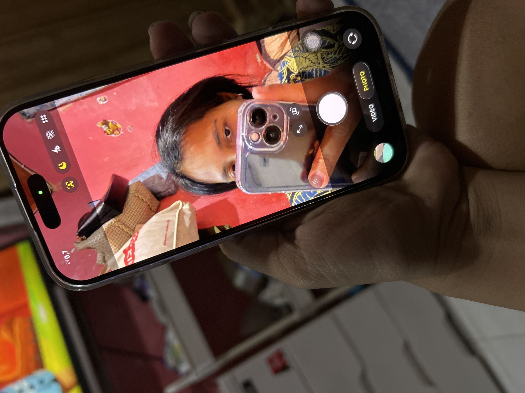 A person's hand holds a smartphone, capturing a selfie. The phone displays the camera interface with options for video and photo. The background appears to be an indoor setting, possibly a room, with furniture and a television screen visible. The phone is held in a way that suggests the person is taking a picture of themselves. The scene is captured in a close-up shot focusing on the phone and the hand holding it.